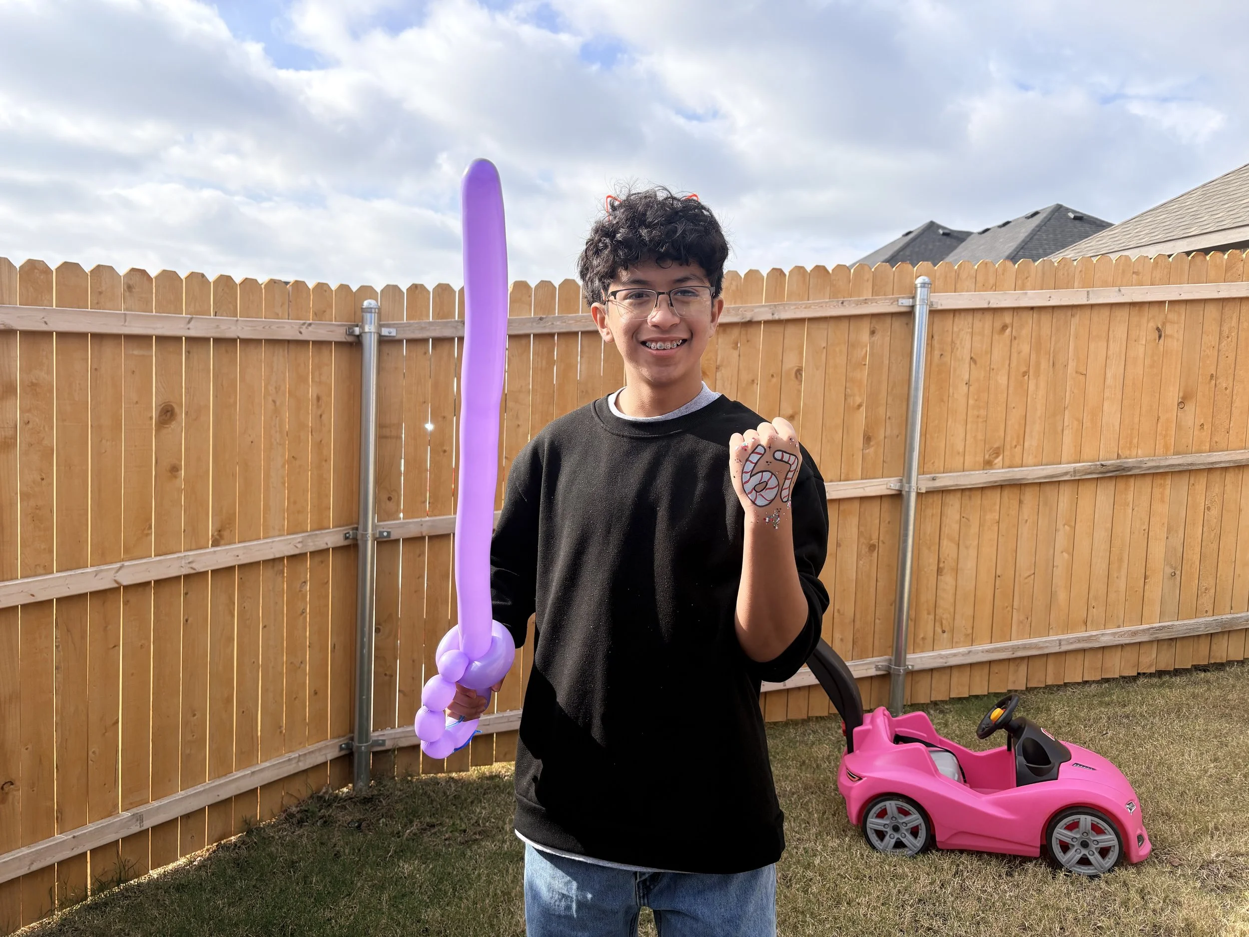 Smiling young boy with glasses holding a purple balloon sword in his right hand and showing a painted lion face on his left hand, standing in a backyard with a pink toy car on the grass behind him and a wooden fence in the background.