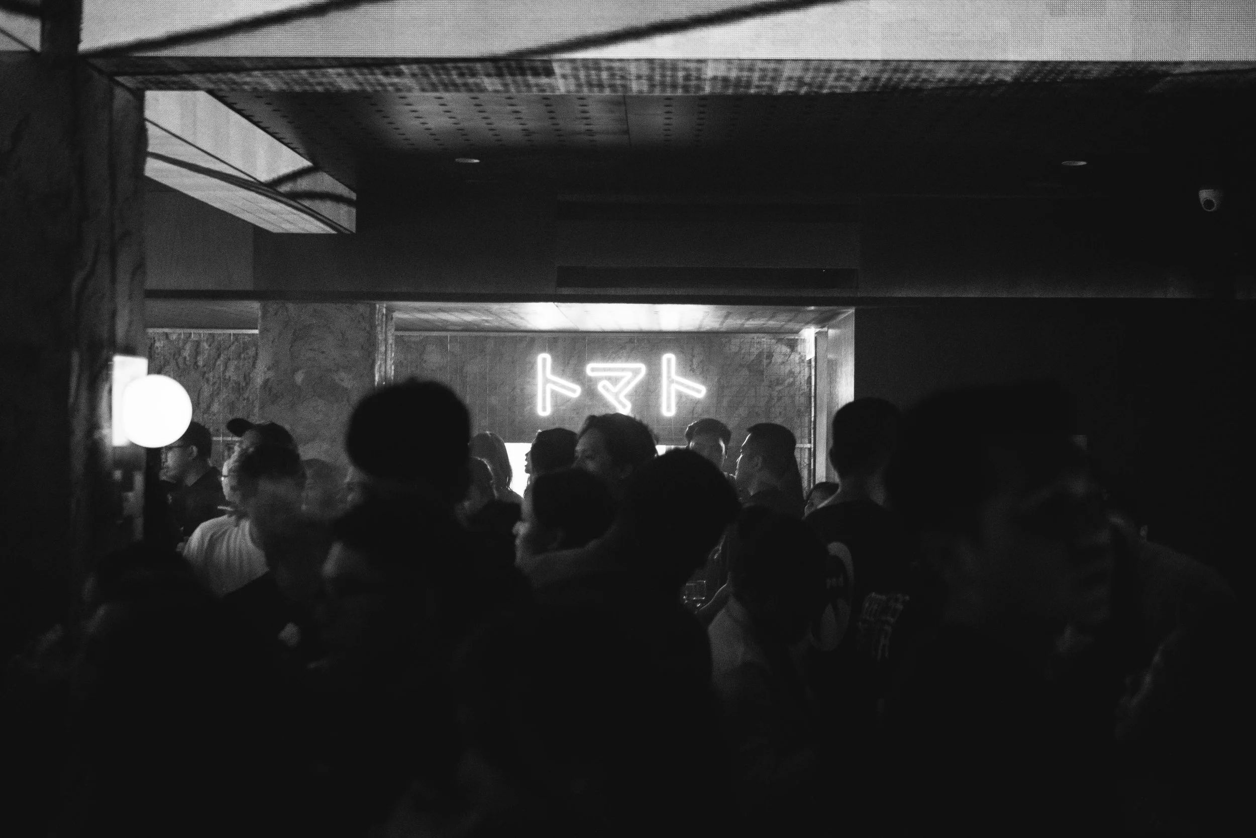 Dimly lit nightclub or bar with a crowd of people gathered, some talking and socializing; neon sign with Asian characters on the wall in the background.