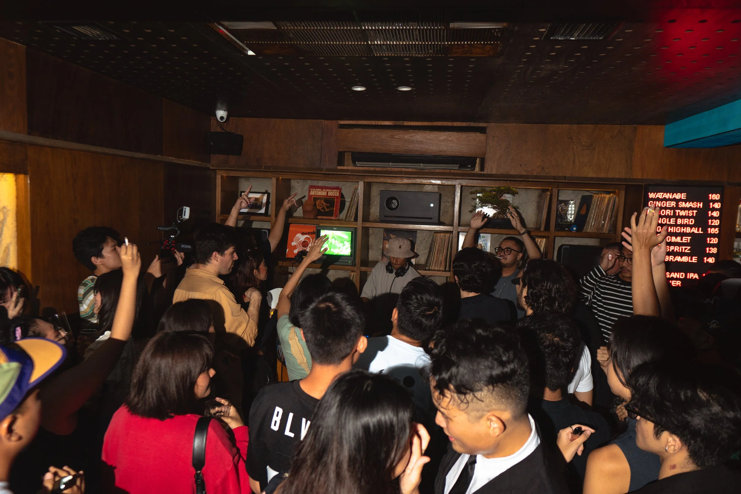 A crowded indoor event with many people standing and raising their hands, in front of a wooden shelf with vinyl records, a small TV, and other items, with a person in the center wearing a hat, possibly DJing or speaking, and a red electronic menu boa
