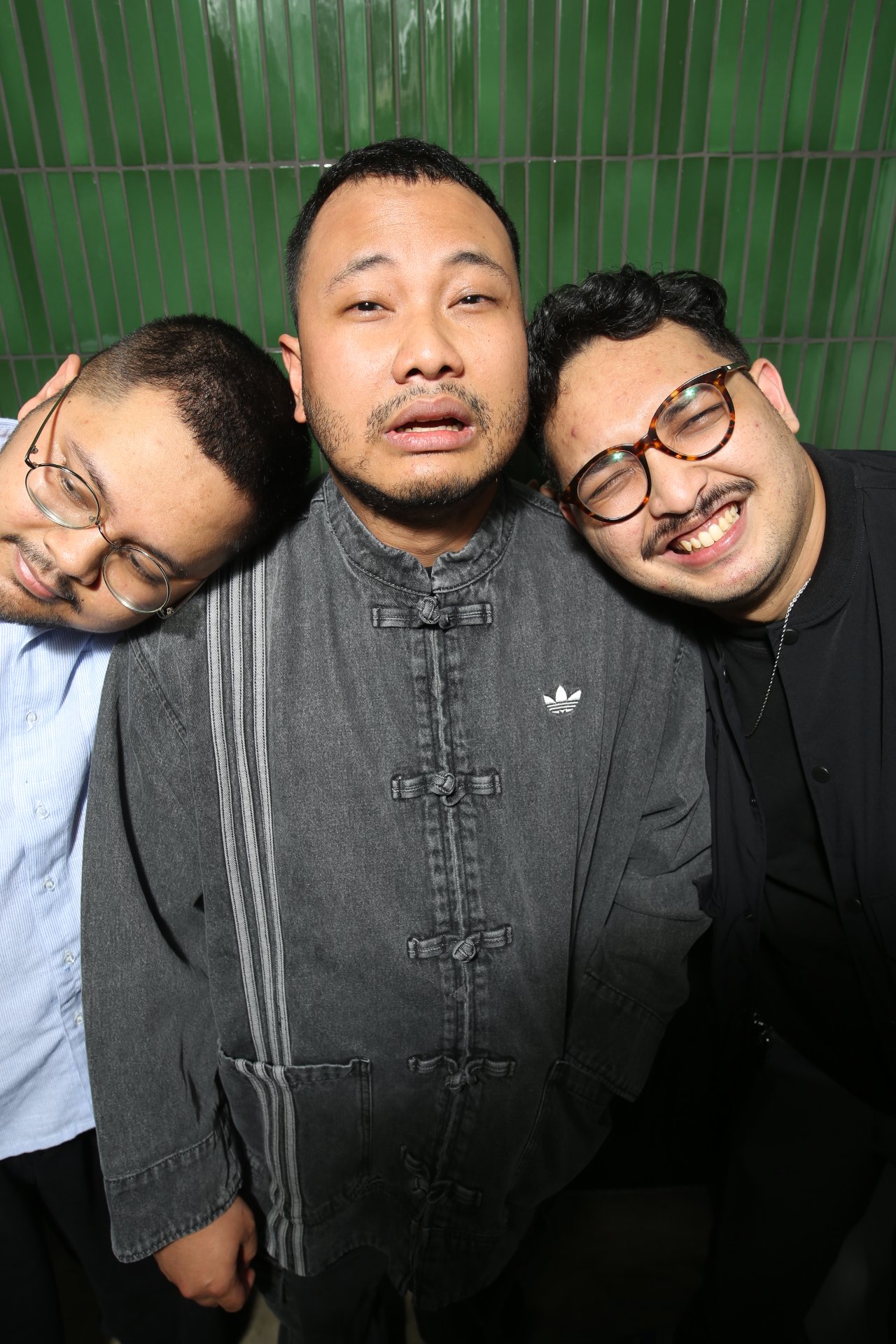 Three men in front of a green tiled background, with the man in the middle wearing a gray jacket with an Adidas logo, and the men on either side wearing glasses and smiling or resting their heads on his.