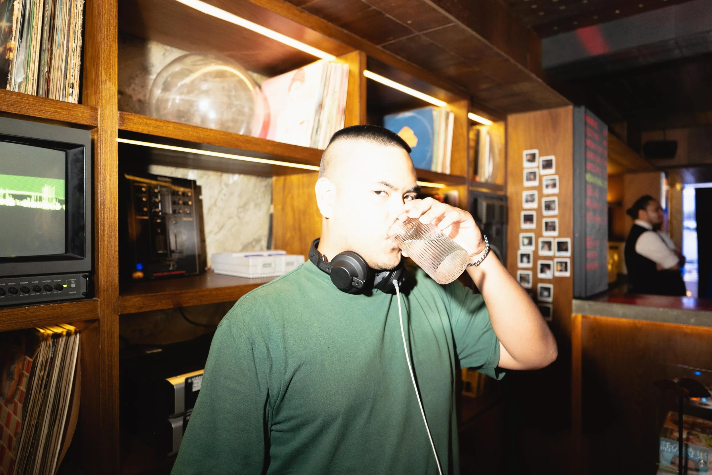 A man with headphones around his neck, wearing a green shirt, is drinking water from a glass in a bar or cafe, with shelves of vinyl records and a Polaroid wall in the background, and a woman in a vest and bow tie visible nearby.