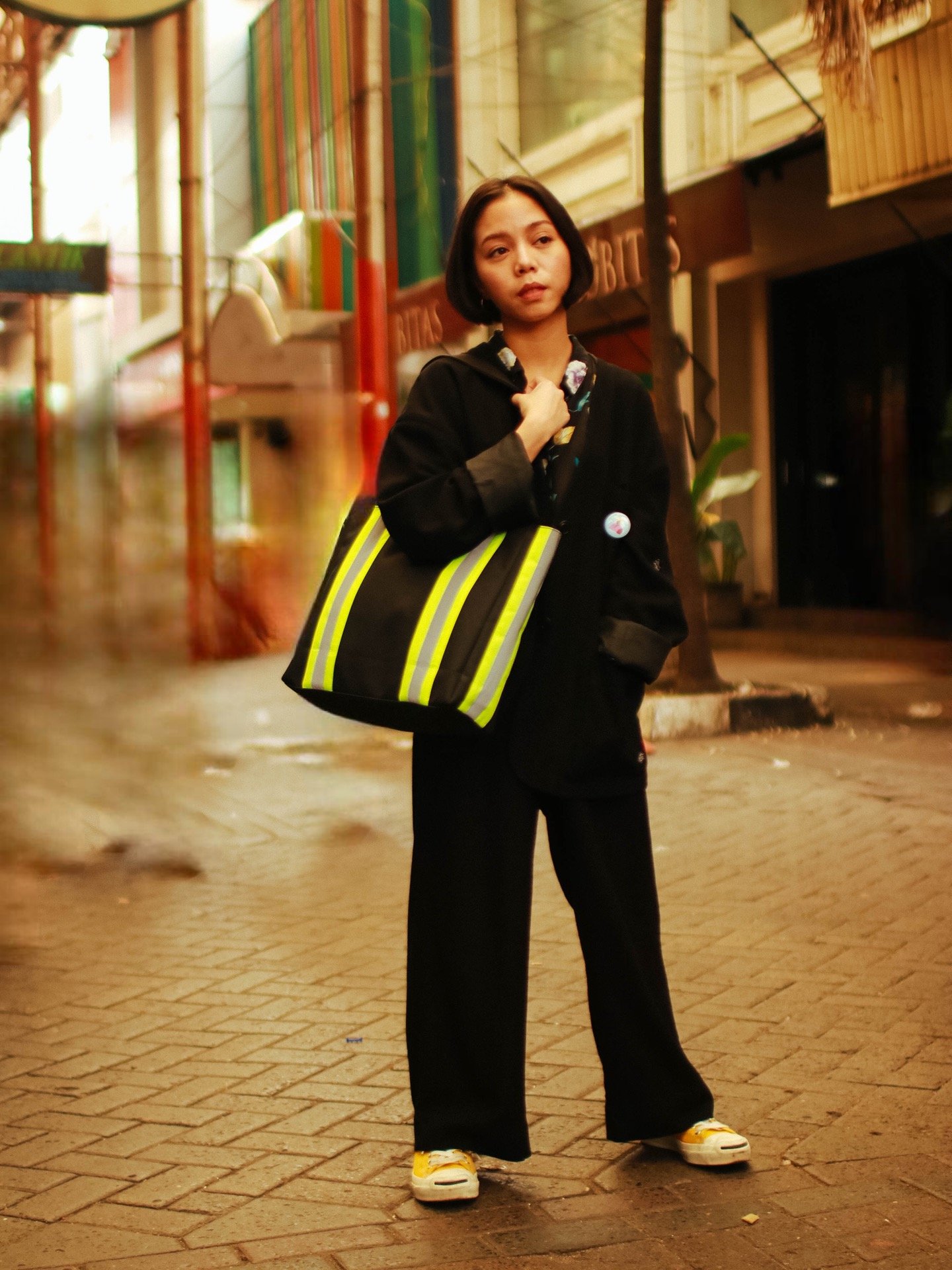 A young woman with short black hair stands on a city street at night, holding a large black bag with yellow reflective stripes over her shoulder. She is dressed in black pants, a black jacket, and yellow sneakers, with a pin on her jacket. The background shows colorful, blurred city buildings and streetlights.