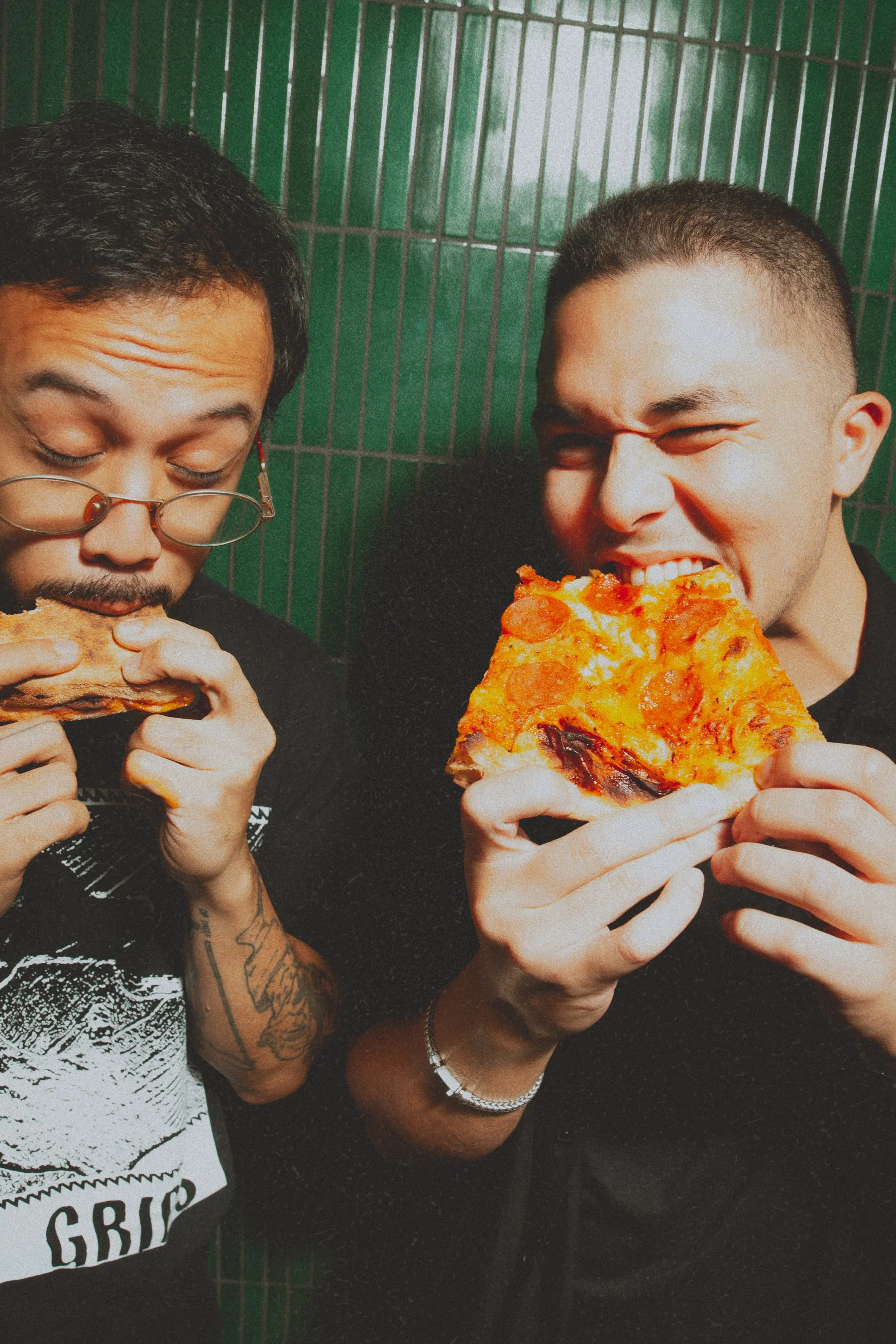 Two men eating pizza in front of a green tile wall. The man on the left has glasses and tattoos, eating a slice of bread, while the man on the right is biting into a large slice of pepperoni pizza, smiling.