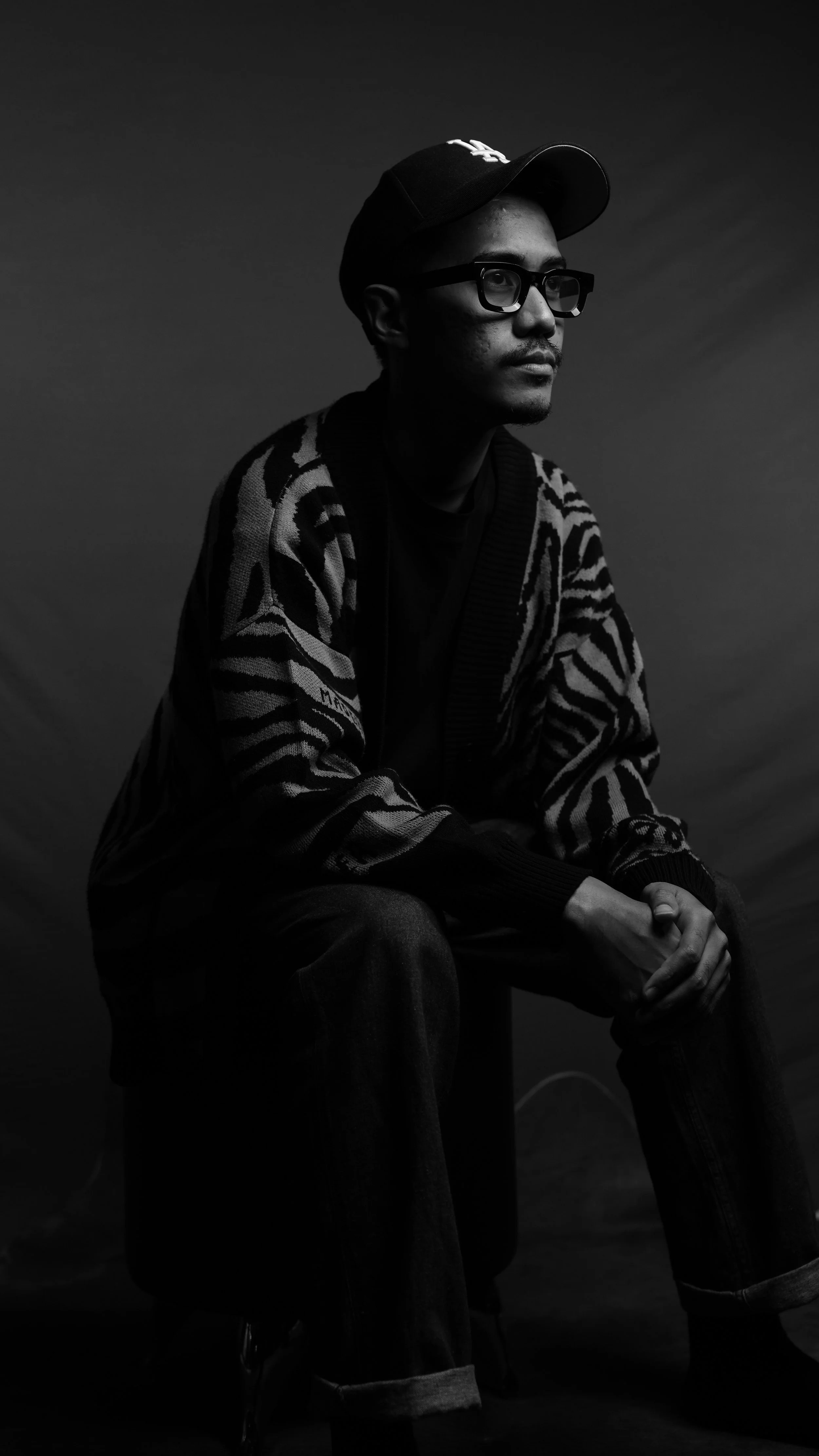 Black and white portrait of a man sitting on a stool, wearing glasses, a black cap, and a patterned jacket with a zebra-like design, looking to the side.