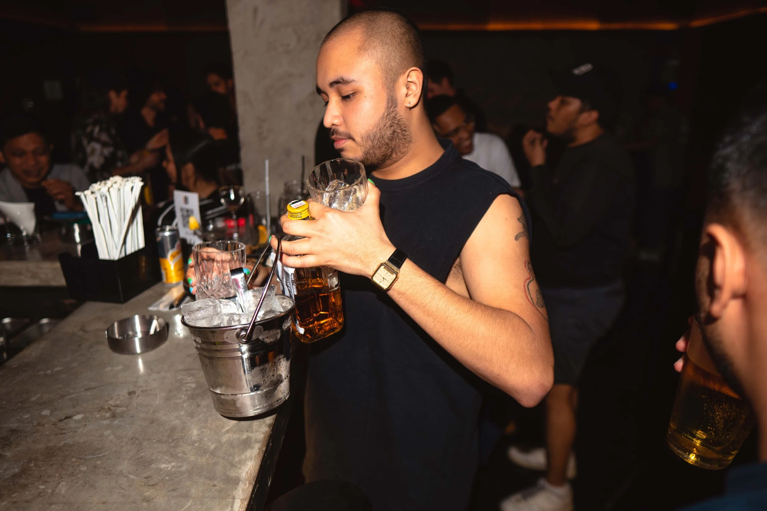 A man in a sleeveless black shirt and a wristwatch is pouring beer from a bottle into a glass in a bar or nightclub. There are other people in the background, some talking and smiling, with bar items and drinks on the counter.