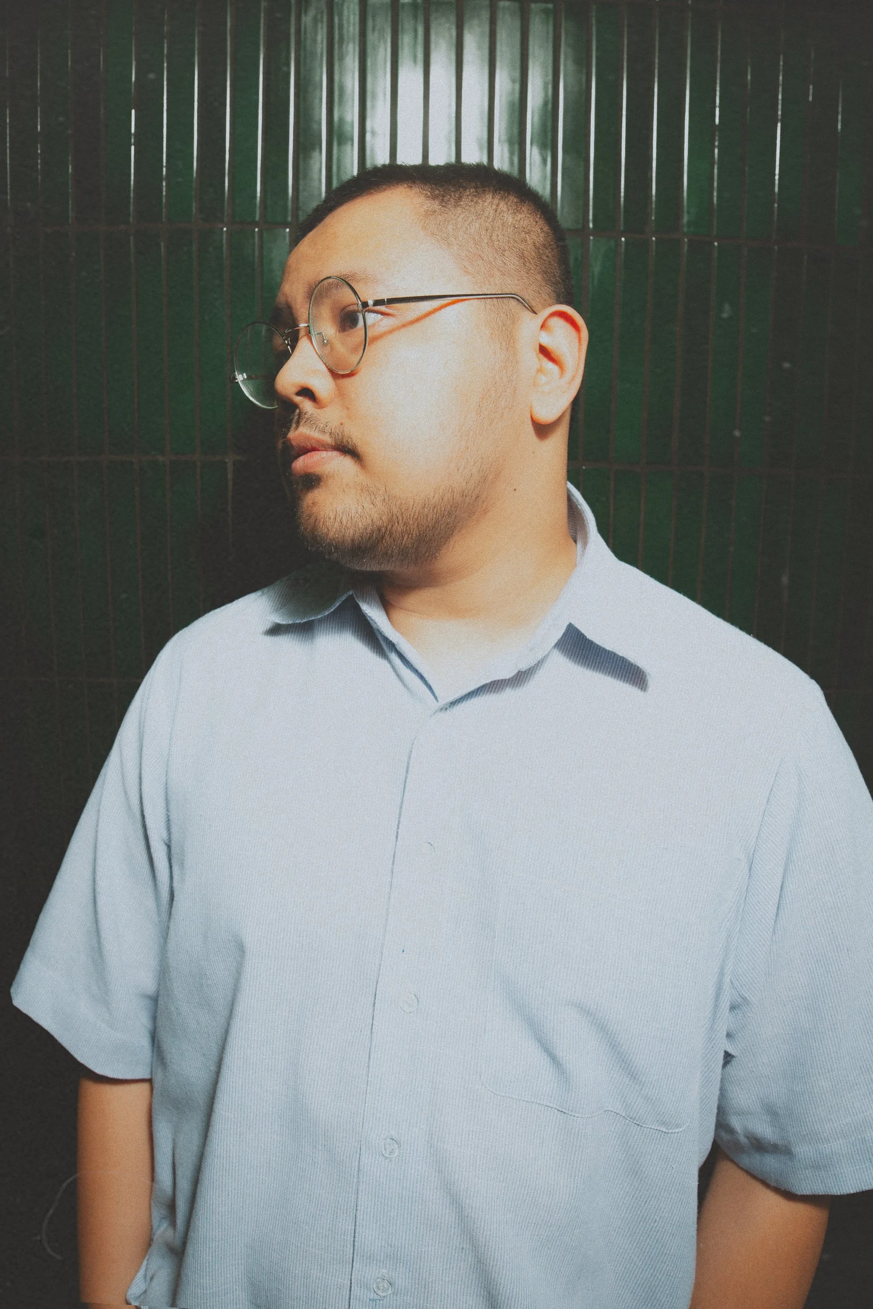A young man with glasses looking to the side, wearing a light blue short-sleeved shirt, standing in front of a dark metal grid background.