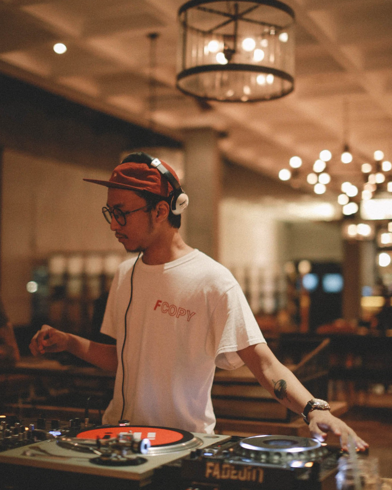 A young man wearing a red baseball cap, glasses, and headphones stands behind DJ equipment in a warmly lit indoor setting, adjusting the controls.