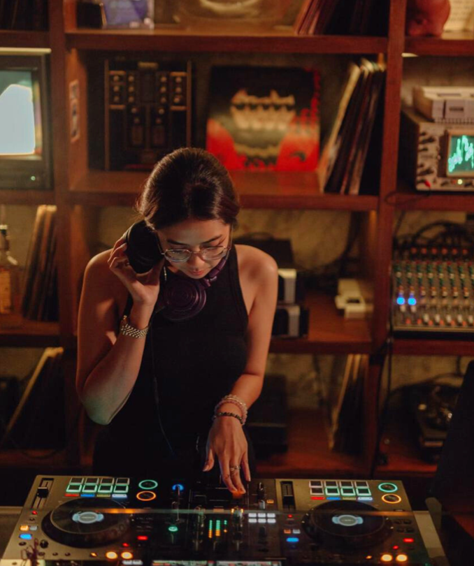 A female DJ with glasses and headphones around her neck, wearing a black sleeveless top, operating a DJ mixer in a cozy room filled with vinyl records, books, and vintage electronics.