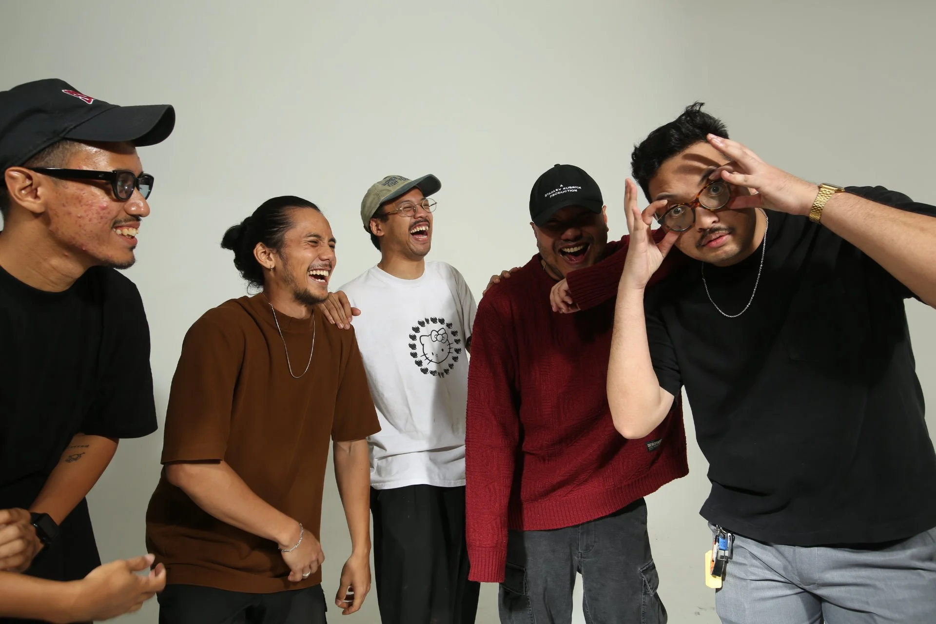 A group of five men laughing and smiling together, with one man adjusting his glasses in front of a plain white background.
