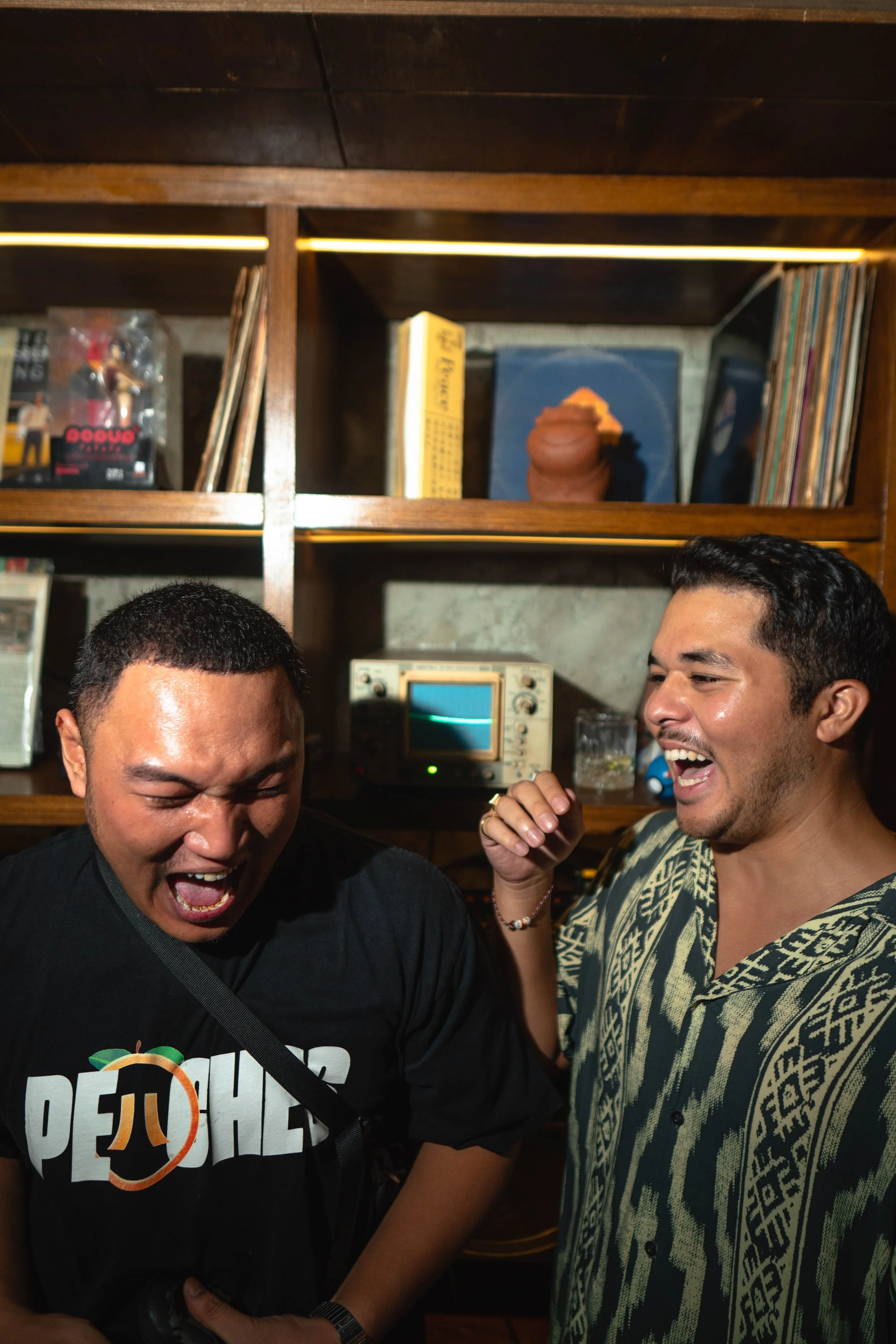 Two men are laughing and celebrating together in a cozy room with a wooden bookshelf behind them, containing vinyl records, books, and vintage items.