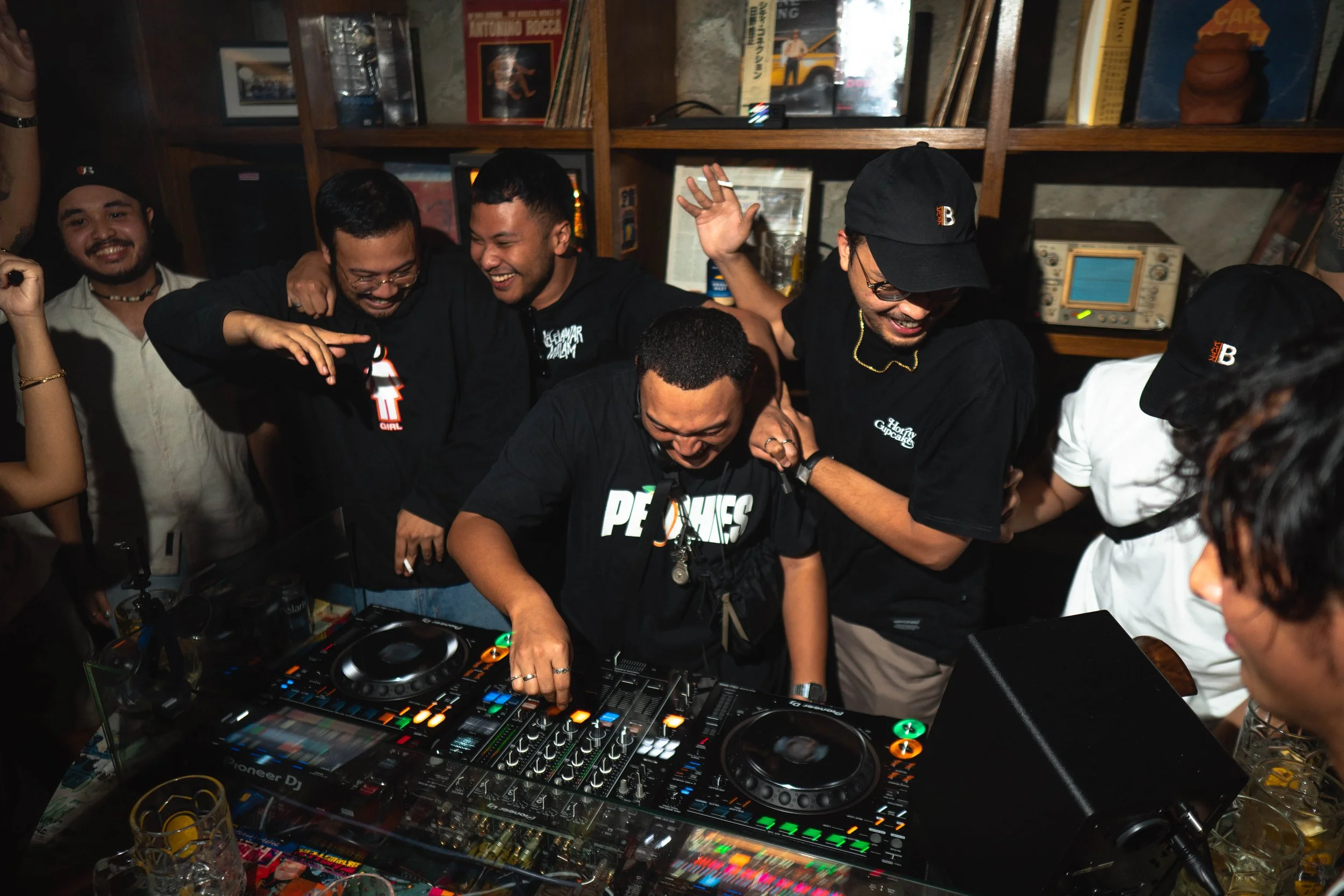 Group of people celebrating around a DJ setup at a party, with some dancing and smiling, in a cozy indoor space with bookshelves and vinyl records in the background.