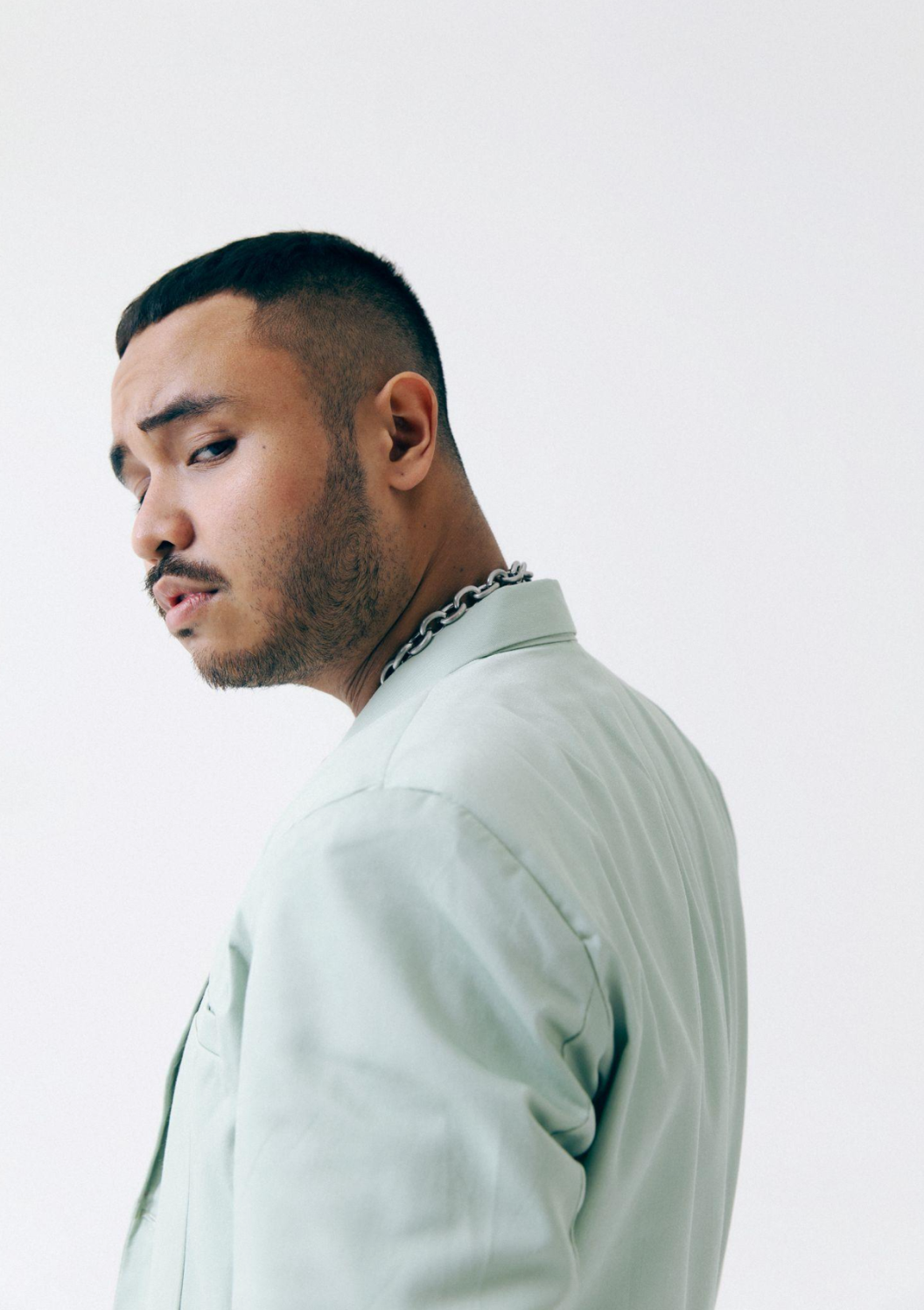 A man with a short haircut, beard, and mustache wearing a light-colored jacket and a silver chain necklace, posing against a plain light background.