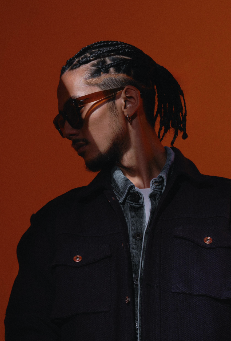 A young man with braided hair and a beard wearing sunglasses, a denim shirt, and a black jacket against an orange background.