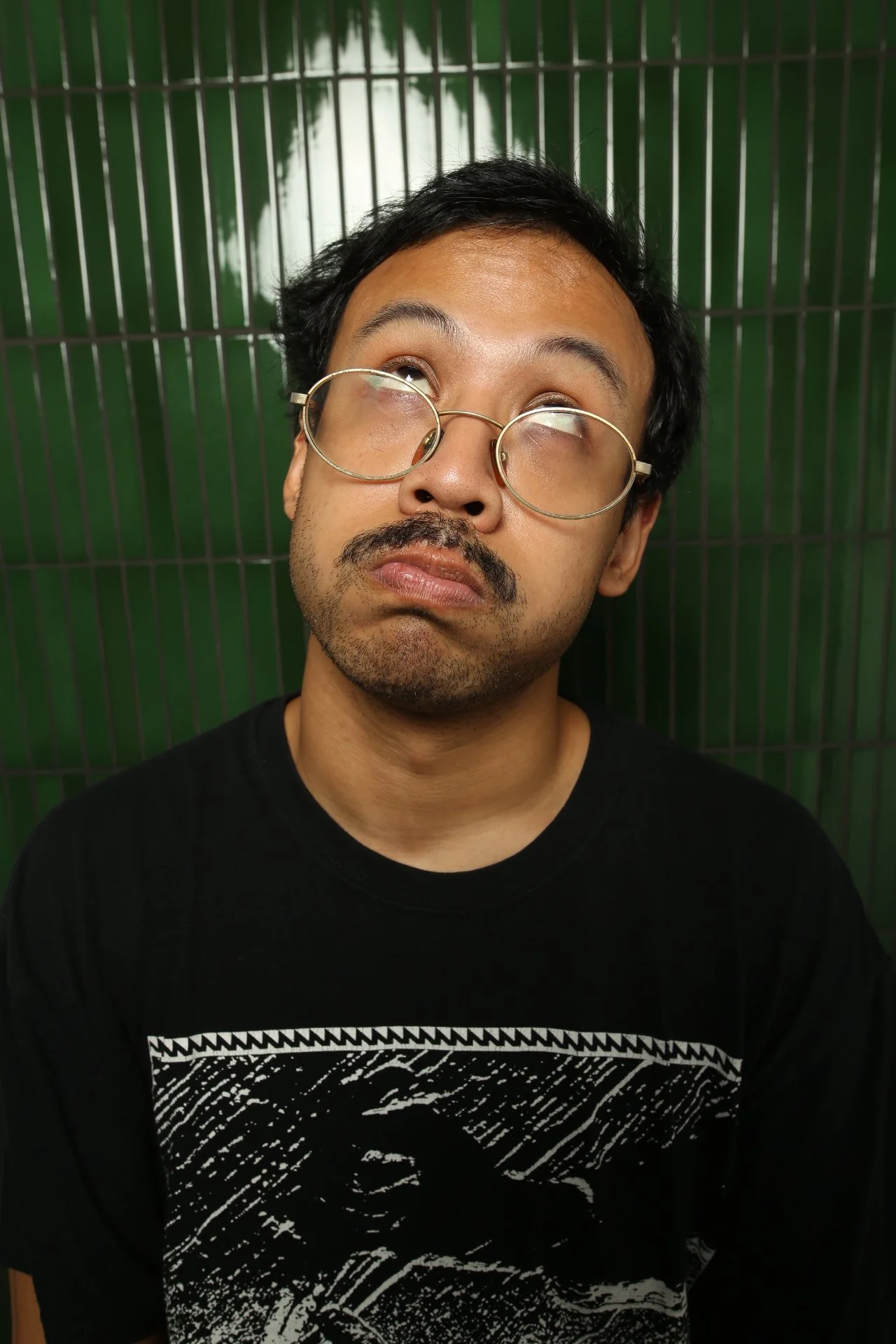 A man with glasses and facial hair looking up with a slightly confused or bored expression, standing in front of a green tiled wall.