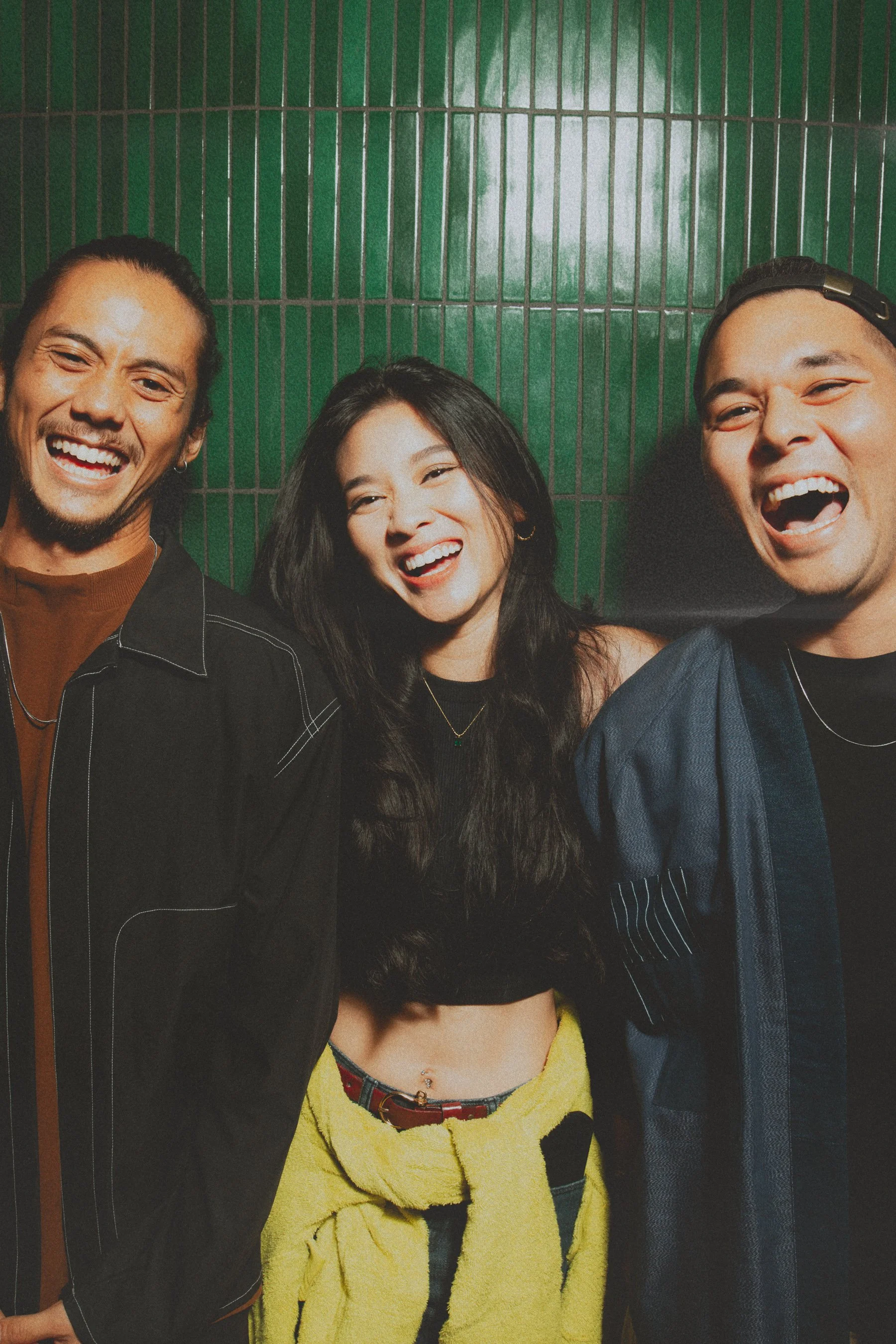 Three friends smiling and laughing together in front of a green tiled wall.