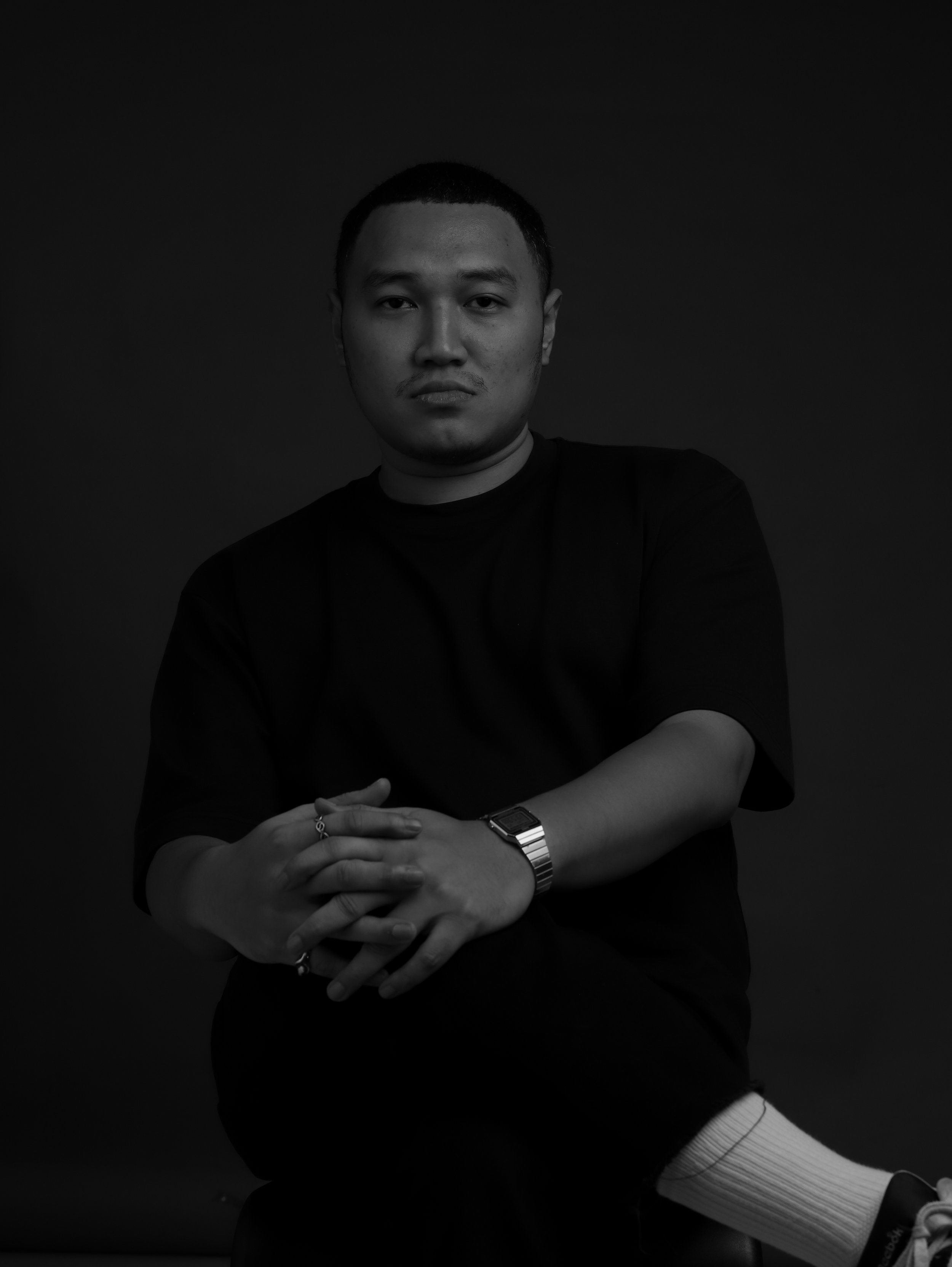 Black and white portrait of a young man sitting with arms crossed, wearing a black T-shirt, a wristwatch, and rings.
