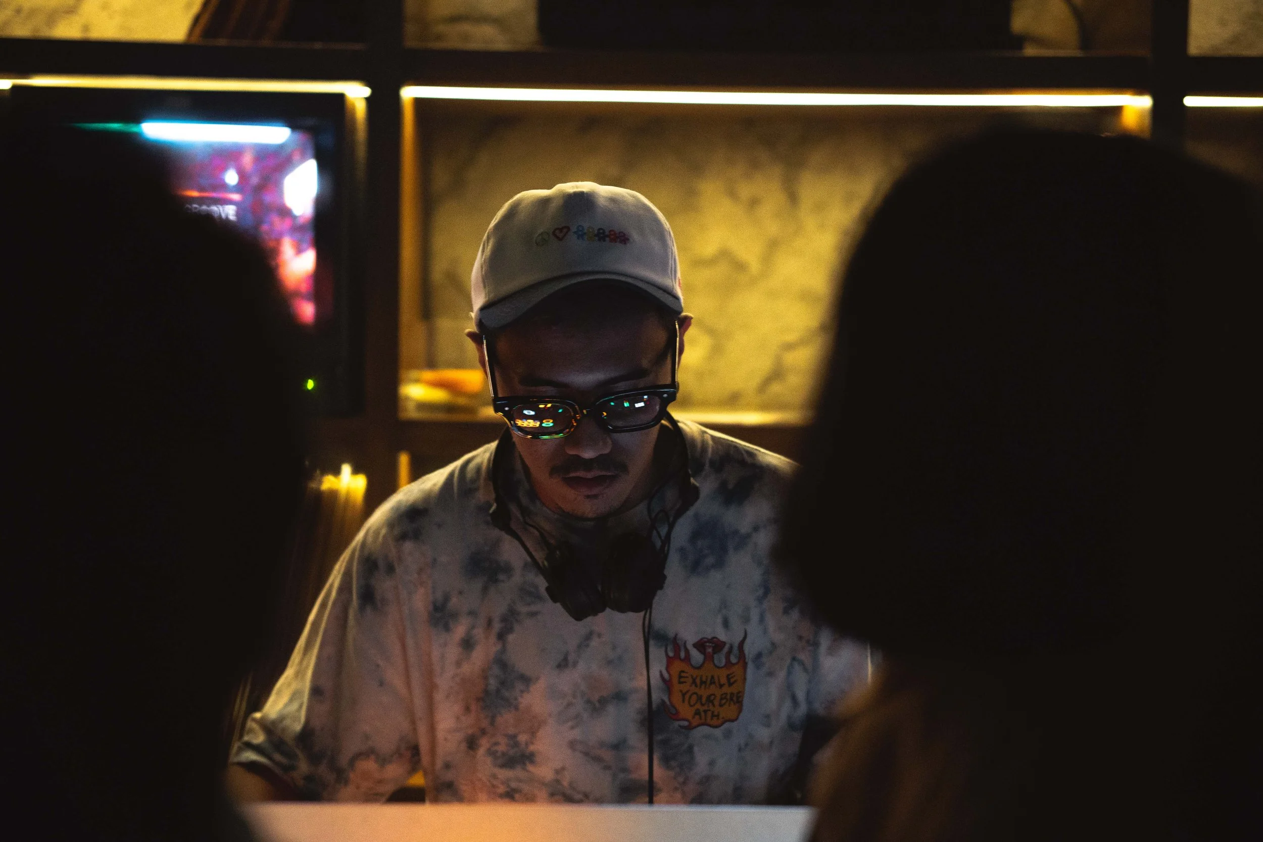 A man wearing a baseball cap, glasses, and a patterned shirt with the phrase "Exhale your breath" on it, is DJing or working on a music setup in a dimly lit environment. There are silhouettes of other people in the foreground.