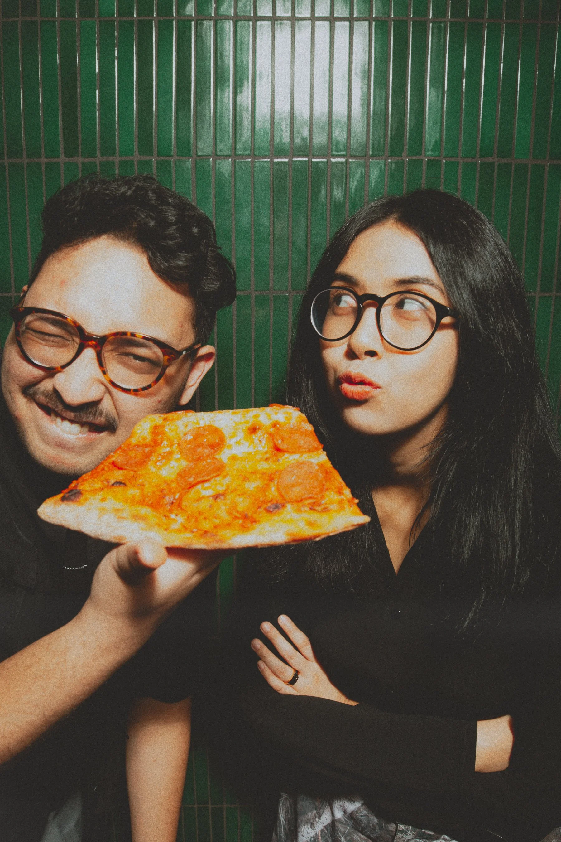 Two people, a man and a woman, pose with a slice of pepperoni pizza against a green tiled wall. The man is smiling with glasses, and the woman is looking at the pizza with a playful expression and glasses.