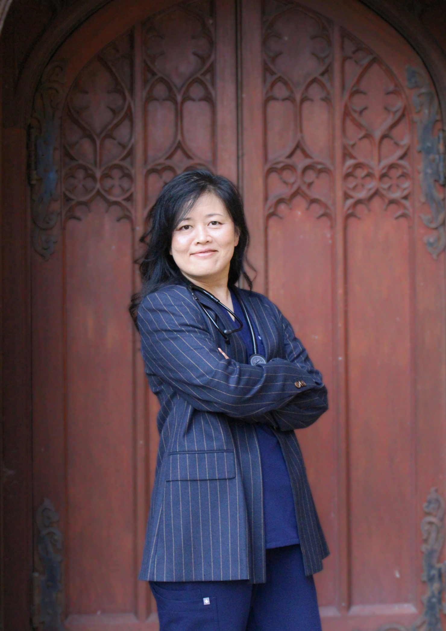 A woman in a pinstripe blazer with a stethoscope around her neck standing confidently in front of a large, ornate wooden door with intricate carvings.