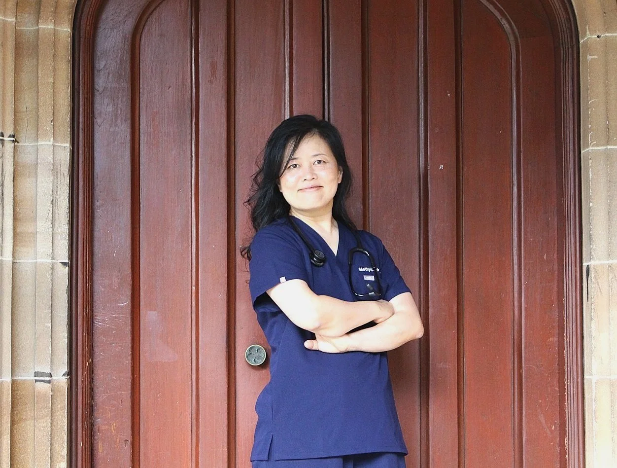 A woman in navy blue medical scrubs standing with arms crossed in front of a wooden door, wearing a stethoscope around her neck.