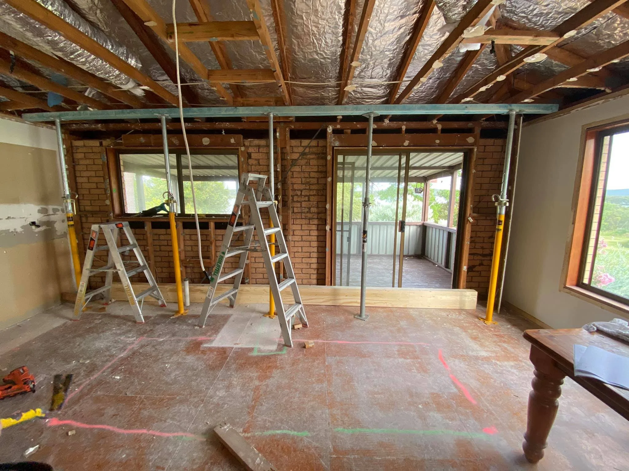 A room under construction with exposed ceiling beams, insulation, and scaffolding. There are ladders, construction tools, and a sliding door leading to an outdoor balcony.