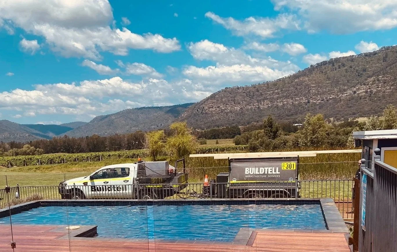 A swimming pool with a wooden deck in the foreground, a black fence, and two construction trucks parked behind it. The trucks have the label 'BUILD TELL INFRASTRUCTURE SERVICES'. In the background, there are green fields, trees, mountains, and a blue sky with fluffy white clouds.