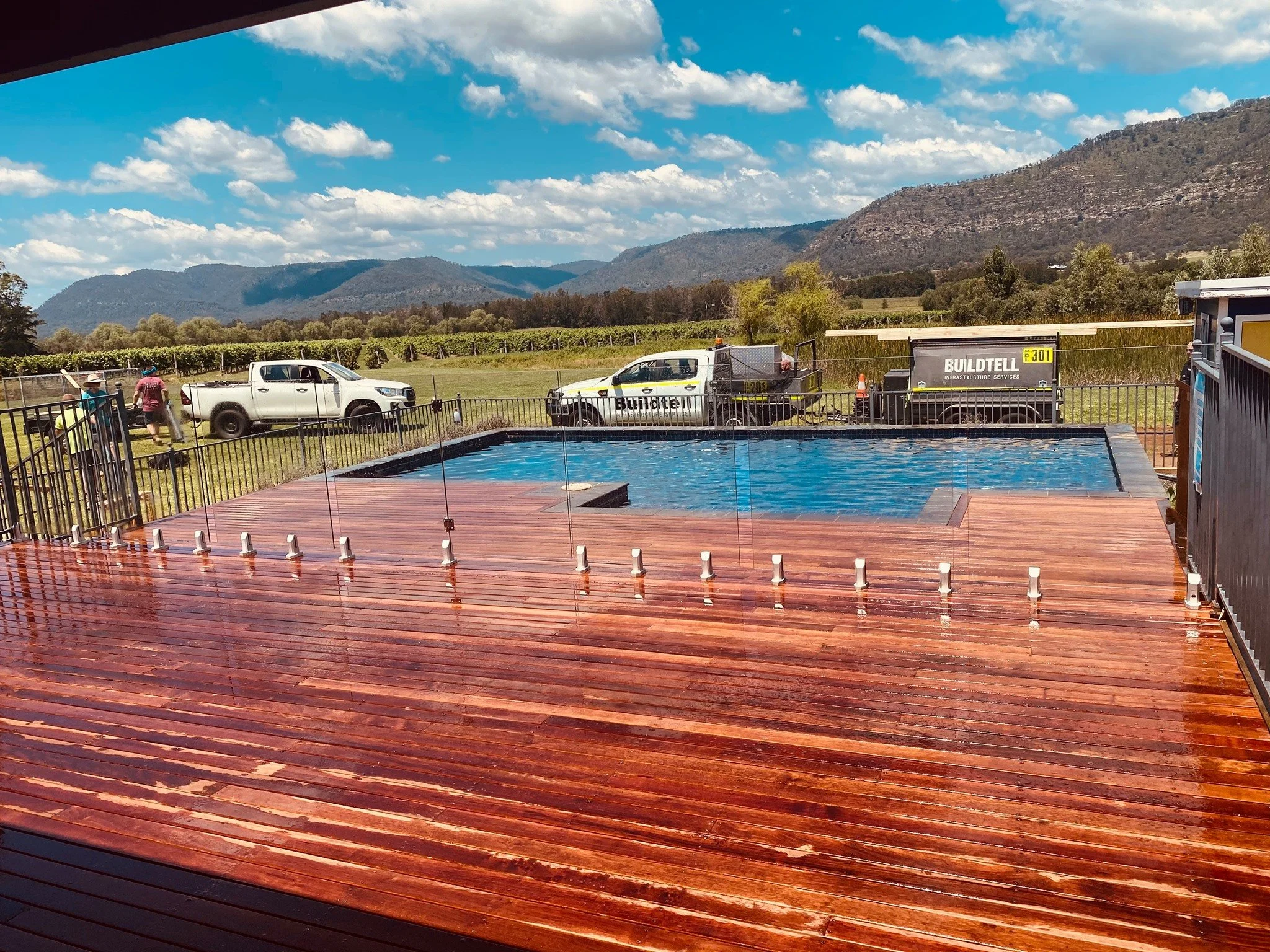 View of a mountain landscape with a vineyard, parked trucks, and a swimming pool in the foreground, under a partly cloudy sky.