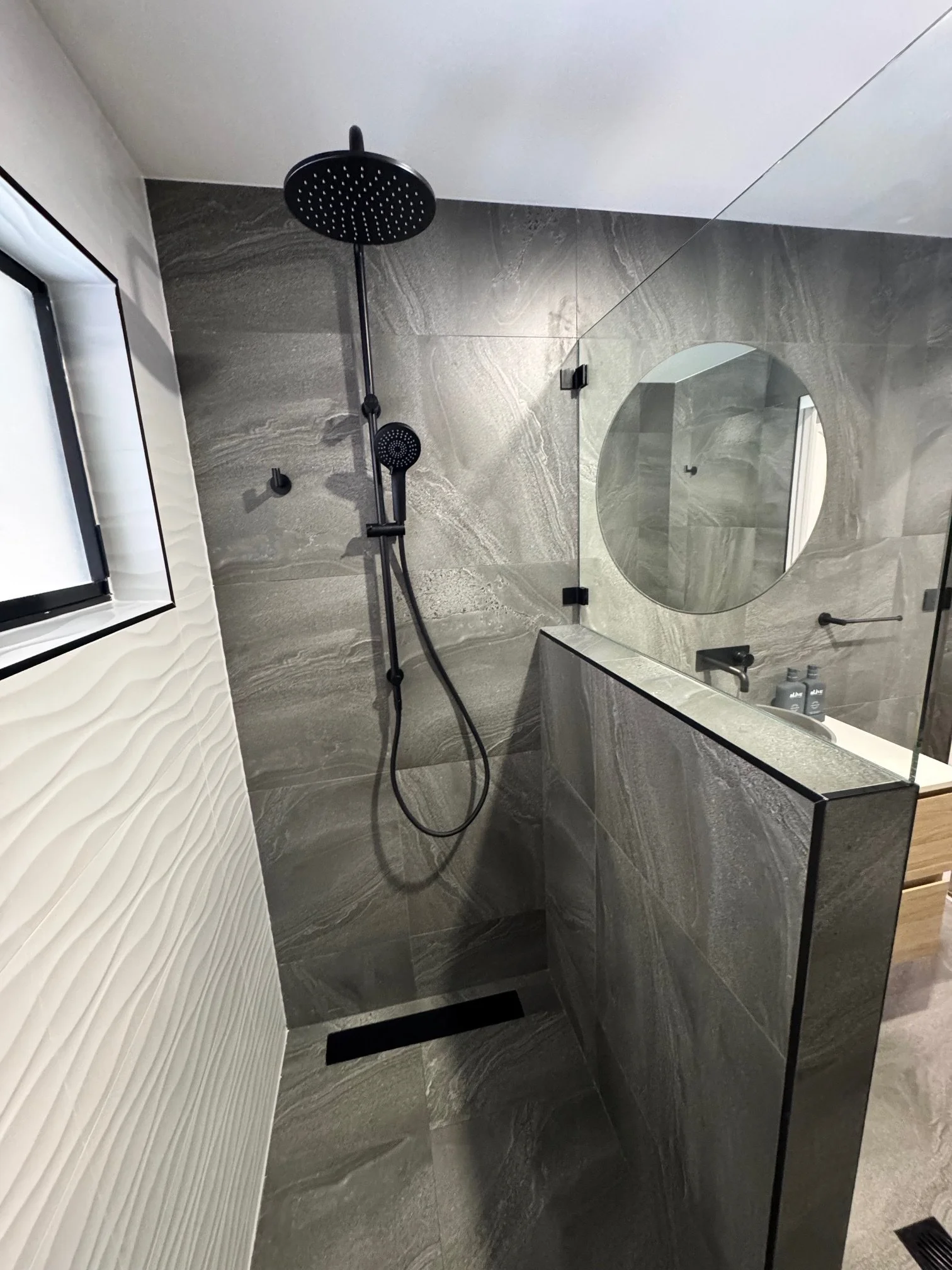 Modern bathroom with a black showerhead and handheld shower in front of large gray marble tiles, round mirror, and a window with textured white wall.