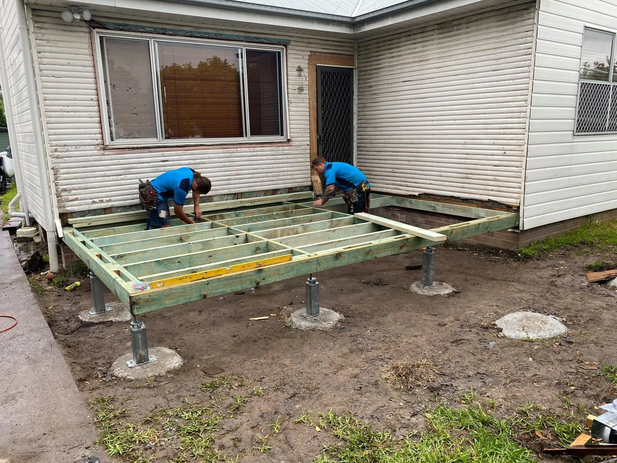 Two workers are building a wooden deck outside of a house. The house has light-colored vinyl siding, and there are large windows and a door with a security screen. The deck frame is partially constructed with greenish wood framing and metal post supports.