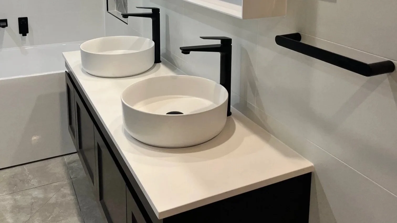 A modern bathroom vanity with two round vessel sinks, black faucets, a mirror, a towel bar, and gray tiled floors.