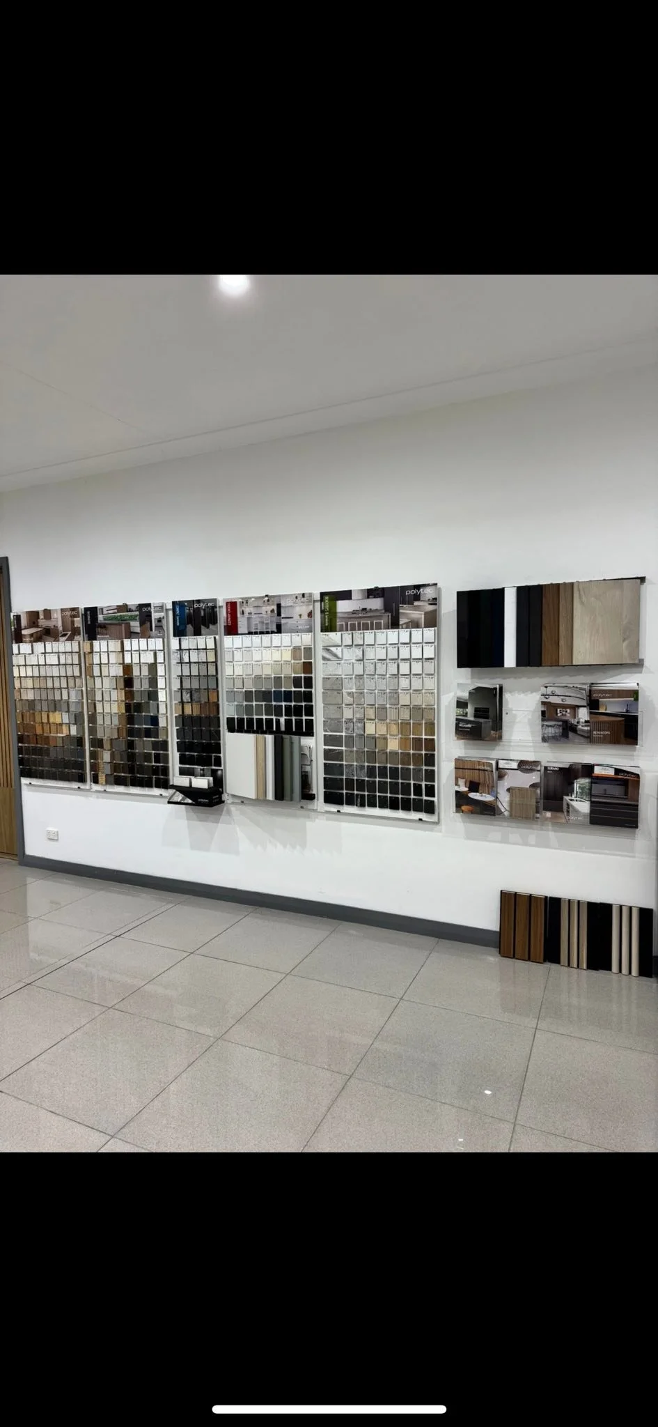 Interior showroom displaying various laminate and wood finish samples on a white wall, with additional product samples and photographs below.
