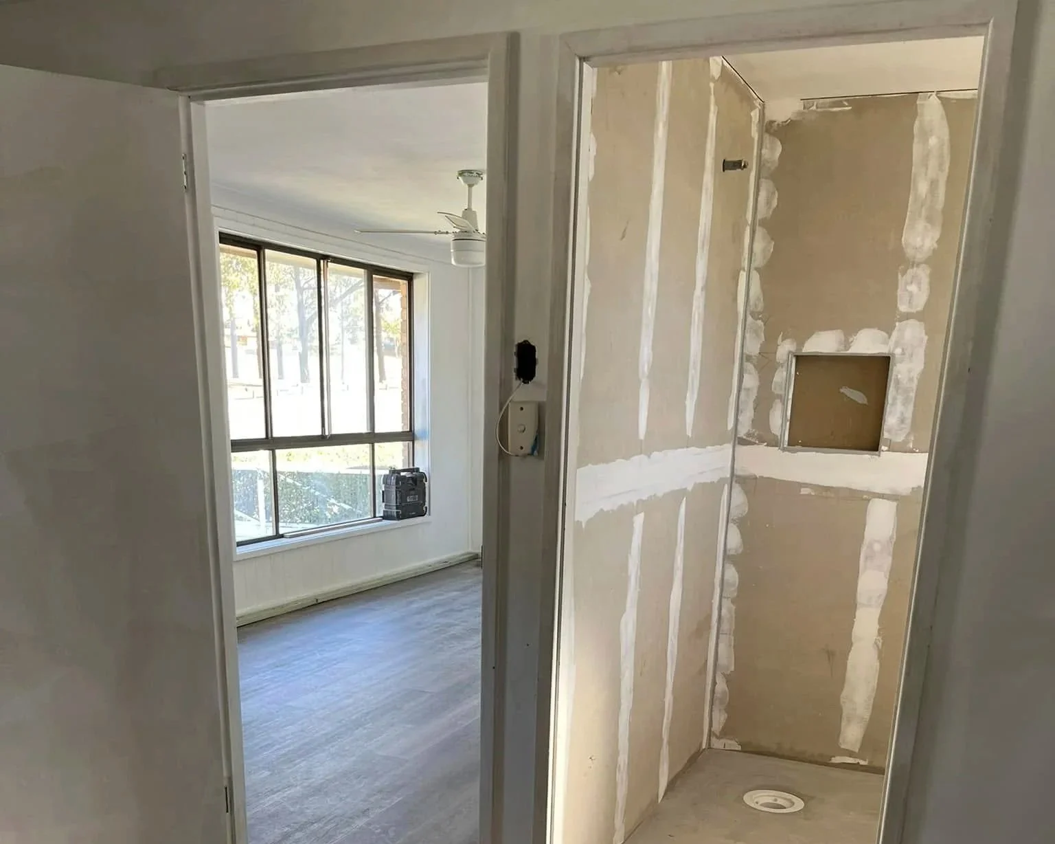 Interior view of a room under renovation with drywall installation, a window with sunlight coming through, and an unfinished shower stall with exposed drywall and piping.
