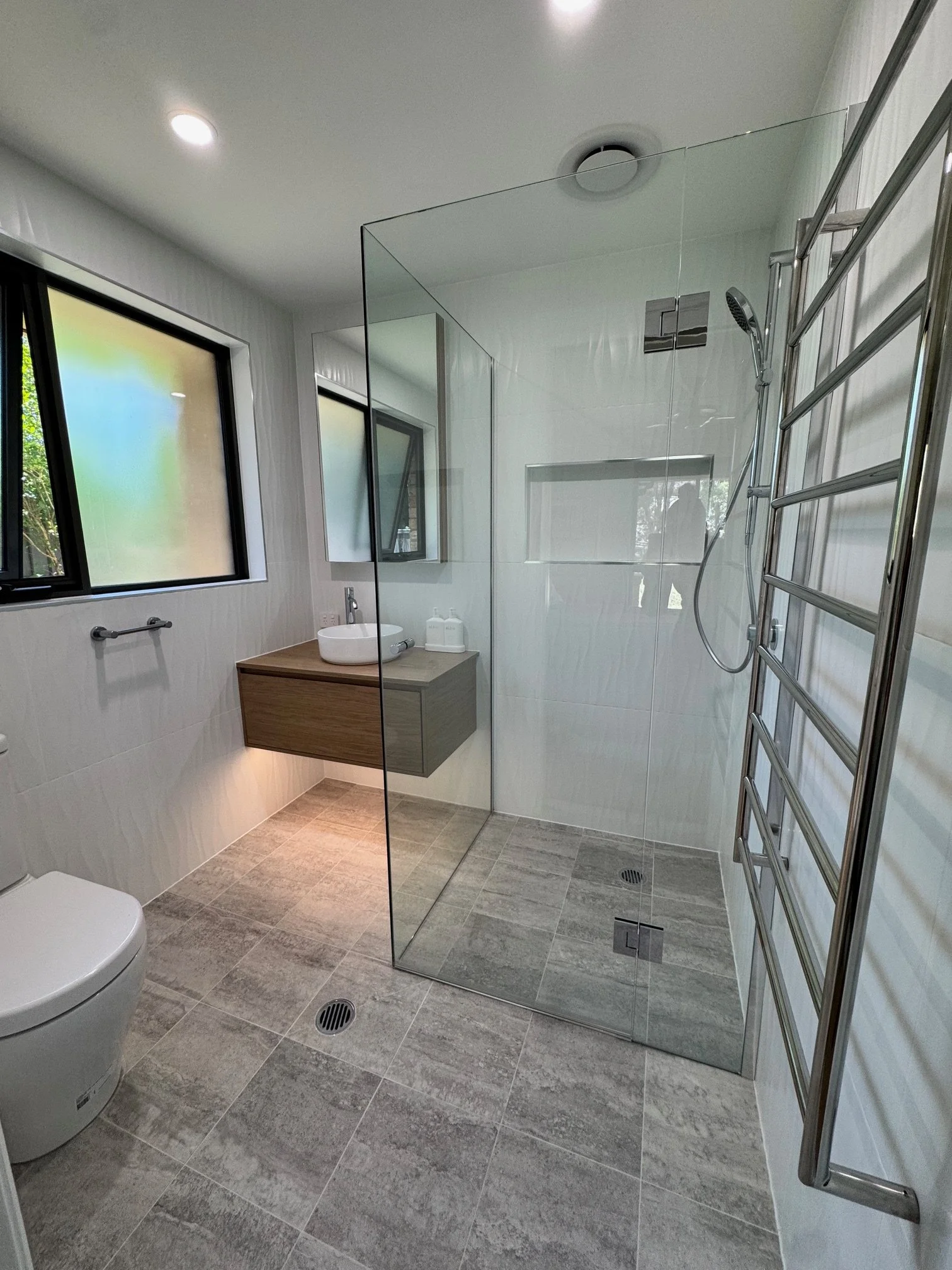 Modern bathroom featuring a walk-in shower with glass panels, a wall-mounted wooden vanity with a vessel sink, a countertop-mounted soap dispenser, a window with tinted glass, a heated towel rail, and beige tile flooring.