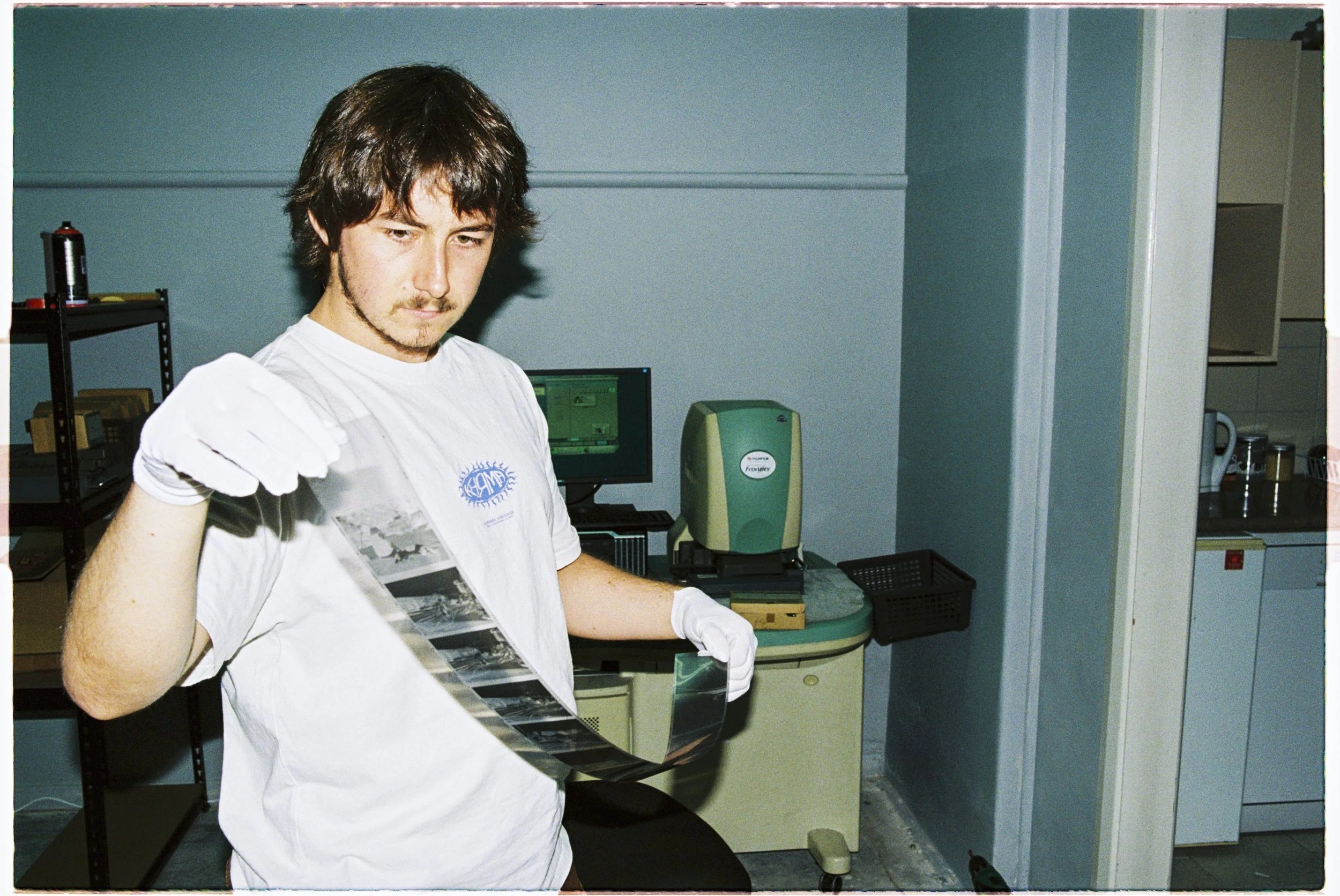 A young man wearing a white t-shirt and white gloves holding a strip of photographic film in a room with office and laboratory equipment.