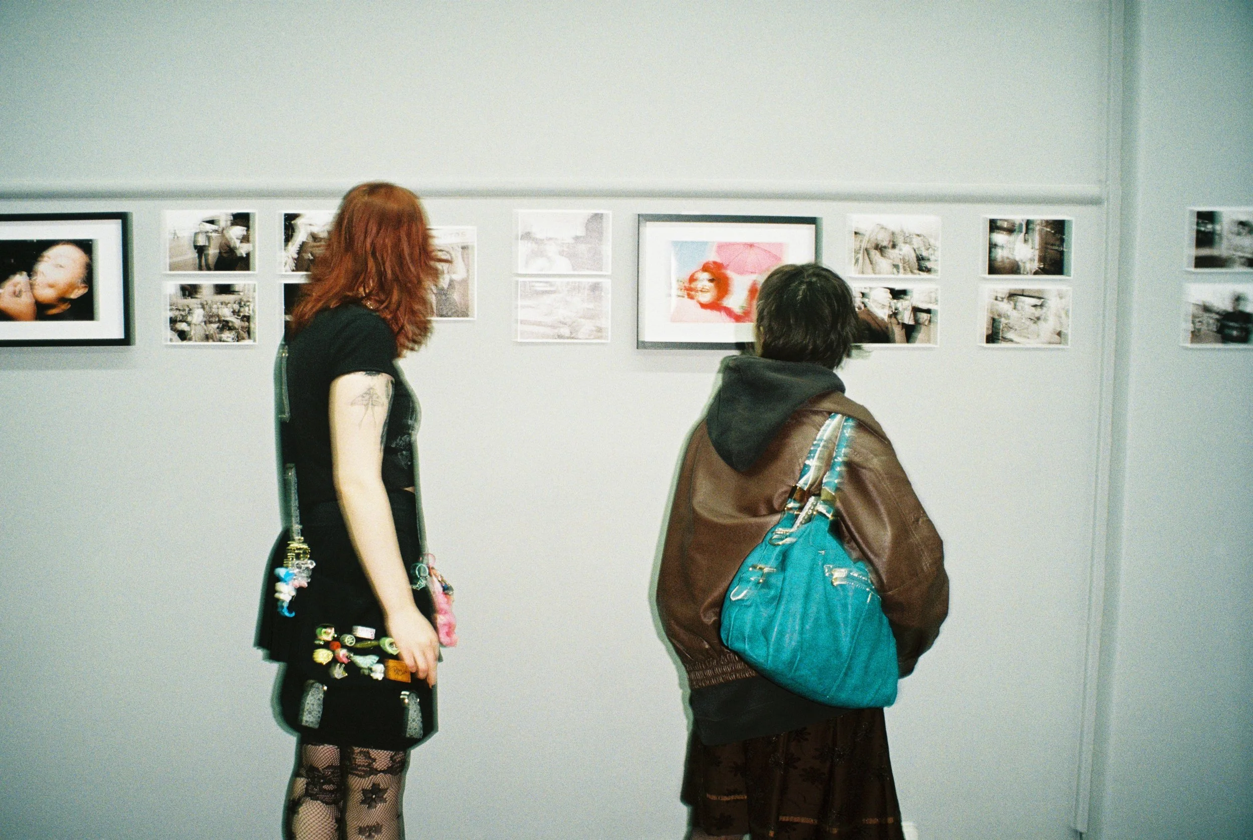 Two people looking at framed photographs on a gallery wall, one with red hair and a black dress with patches, and the other with dark hair, a brown jacket, and a large blue bag.