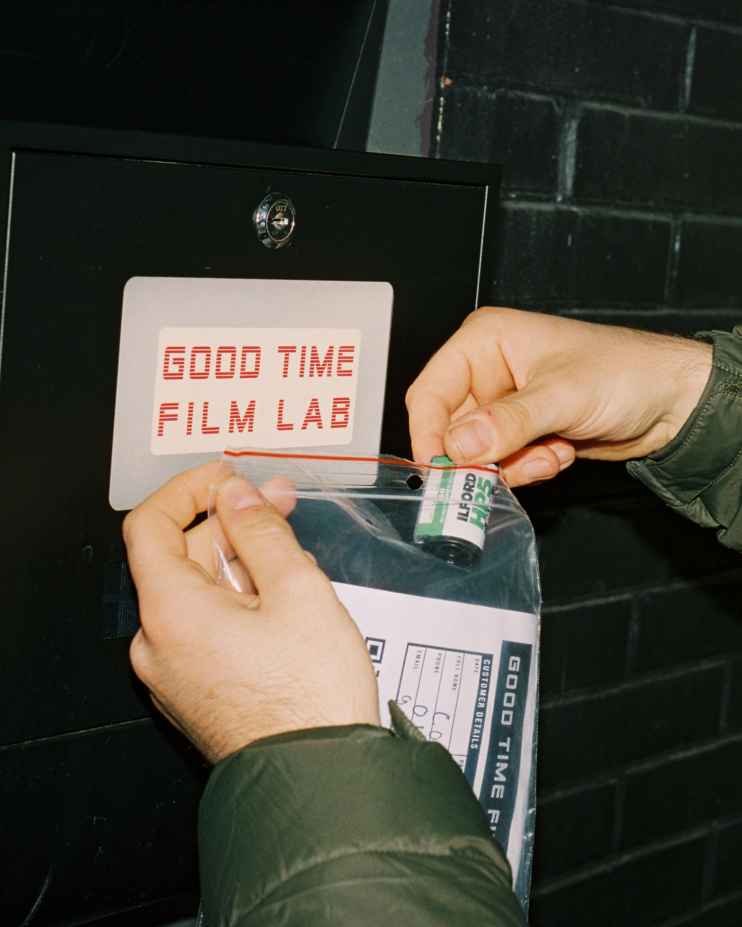 Person inserting film into a vending machine labeled 'Good Time Film Lab' with a sign that reads 'Good Time Film Lab' inside the machine.