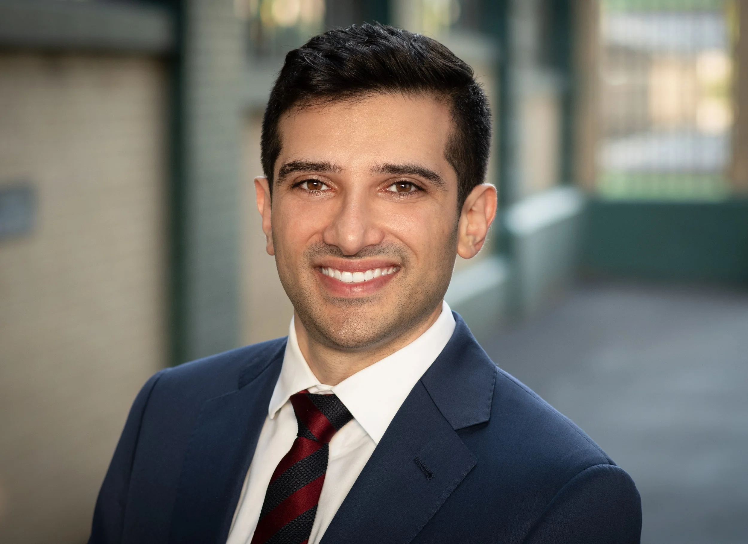 Professional headshot of Dr. Peter Bittar, a man with short dark hair wearing a navy blue suit, white shirt, and red striped tie, smiling in an outdoor urban setting with a blurred background.