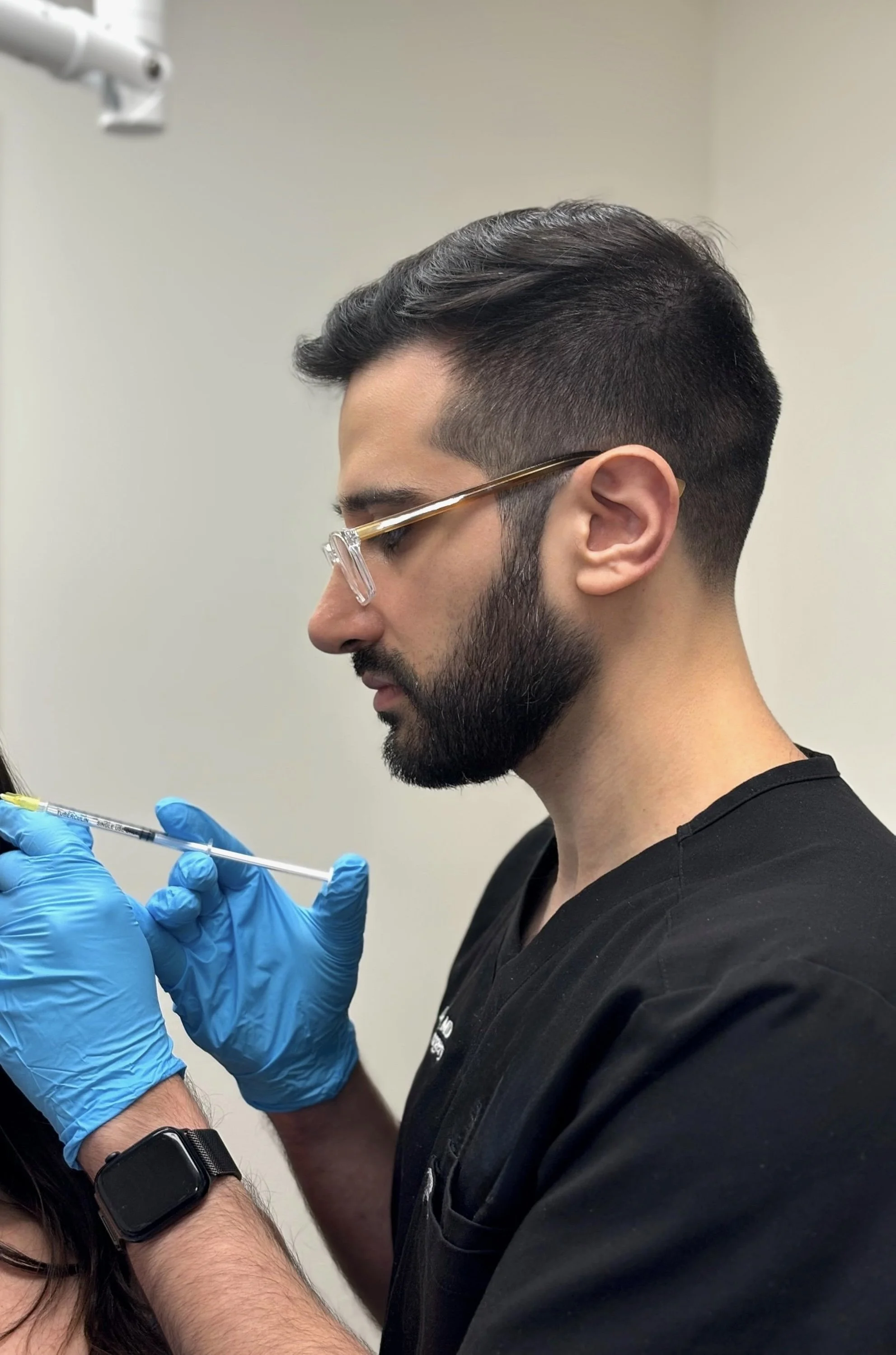 The side profile of Dr. Peter Bittar, a man wearing glasses and black scrubs, carefully holding a syringe while wearing blue medical gloves, preparing for an injection in a clinical setting.