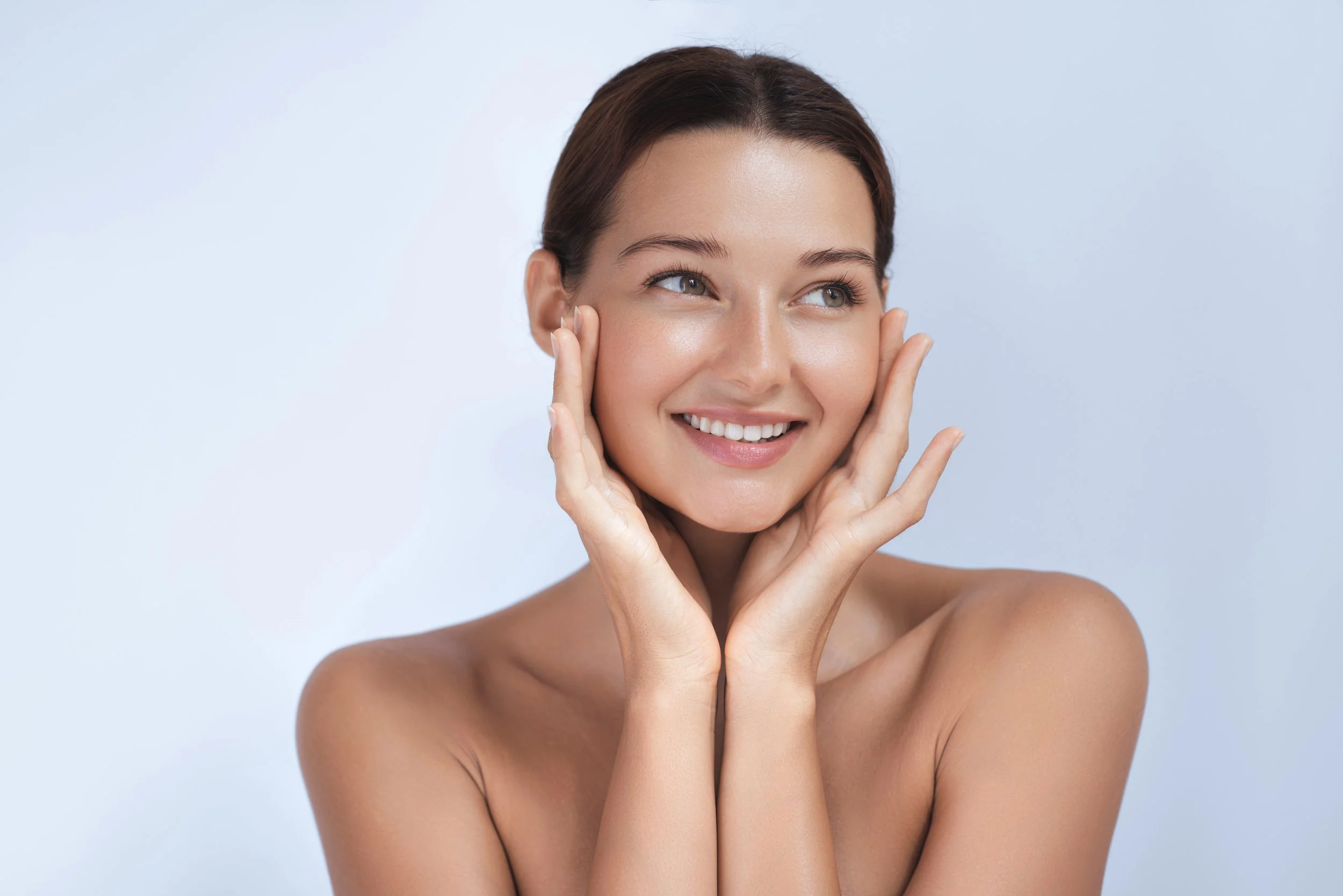 Close-up of a smiling woman with glowing skin and hair pulled back, gently touching her face with both hands against a light blue background.