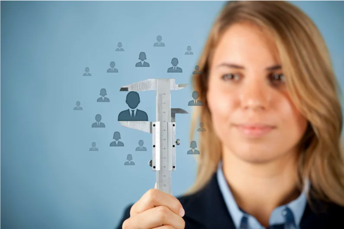 A woman holds a caliper around a cutout of a person's silhouette, with multiple other silhouettes in the background, representing measurement or analysis of a group or individual.