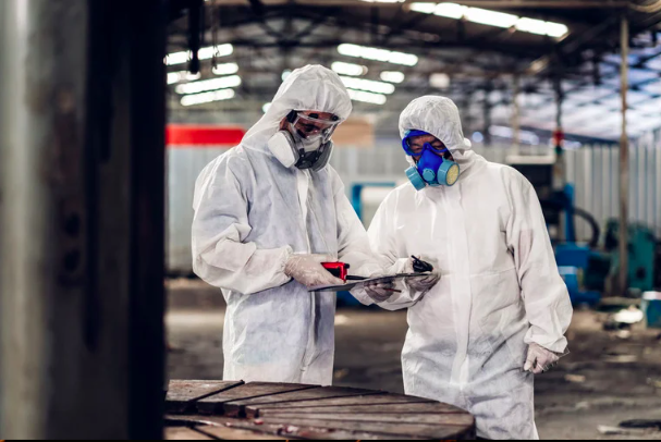 Two workers in protective suits and masks inspecting a clipboard inside an industrial warehouse.