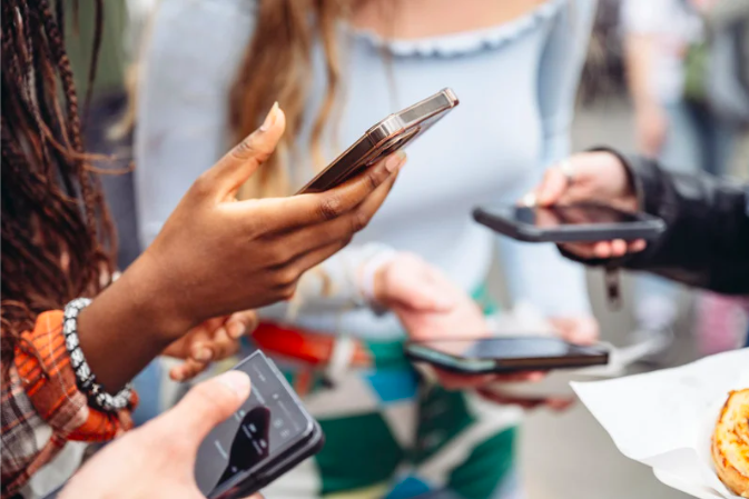 Multiple people gathered, looking at their smartphones, with a partially visible food item (possibly a pie or pastry) on the right.