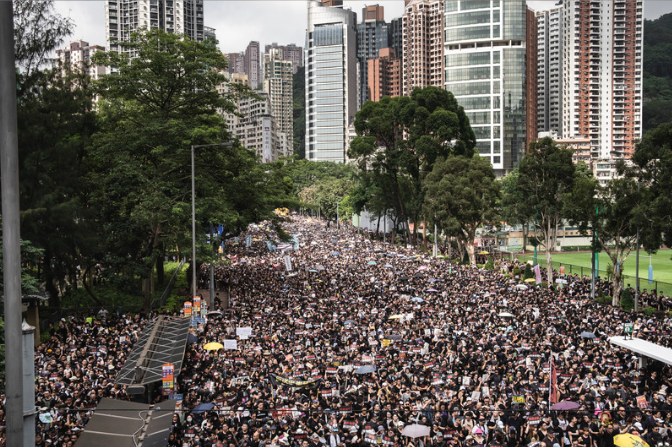 Large crowd of people protesting in an urban park with tall buildings and trees in the background.