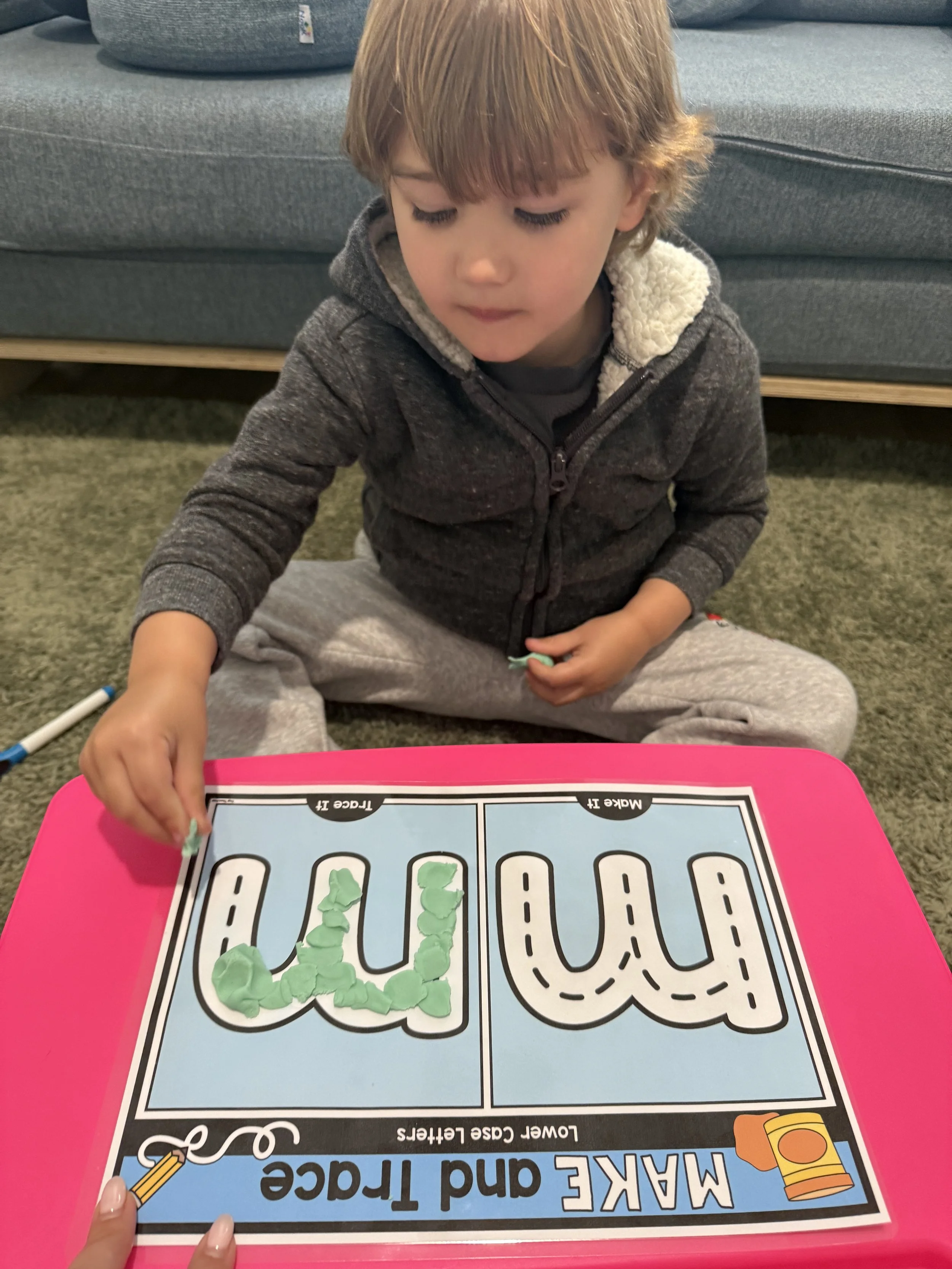 A young boy practicing writing on a maze and trace activity sheet with letter 'U' at a pink table.