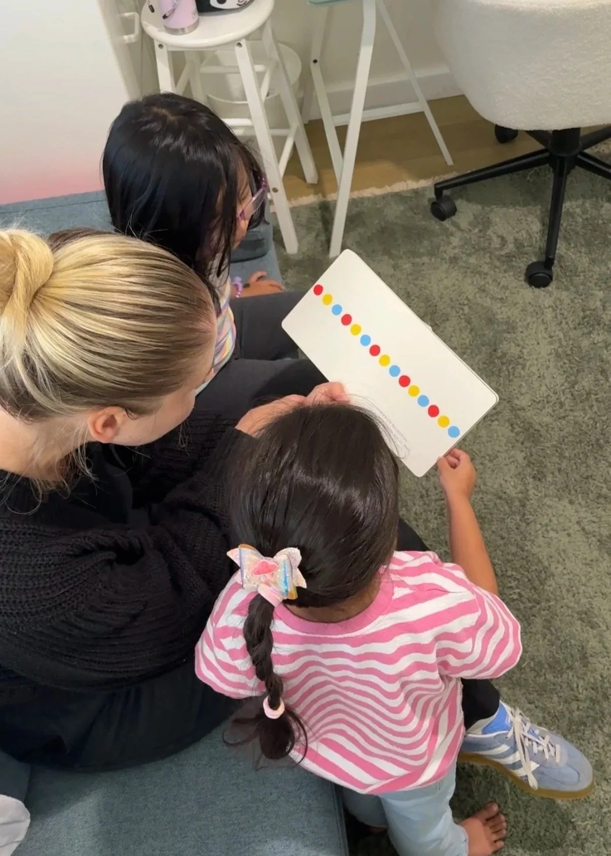 Group of children and an adult sitting on the floor, looking at a whiteboard with a pattern of colored dots, in a cozy room with a green carpet.