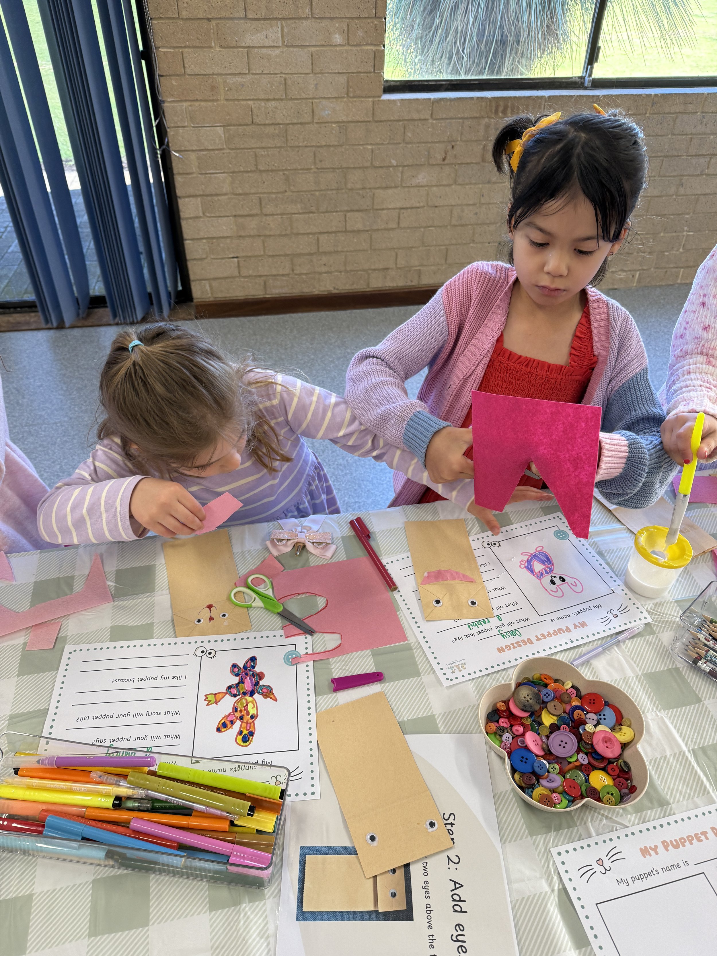 Two young girls are crafting with felt, buttons, and markers at a table filled with crafting supplies, including colorful pens, scissors, and paper puppets. They are creating puppet designs with paper and felt.