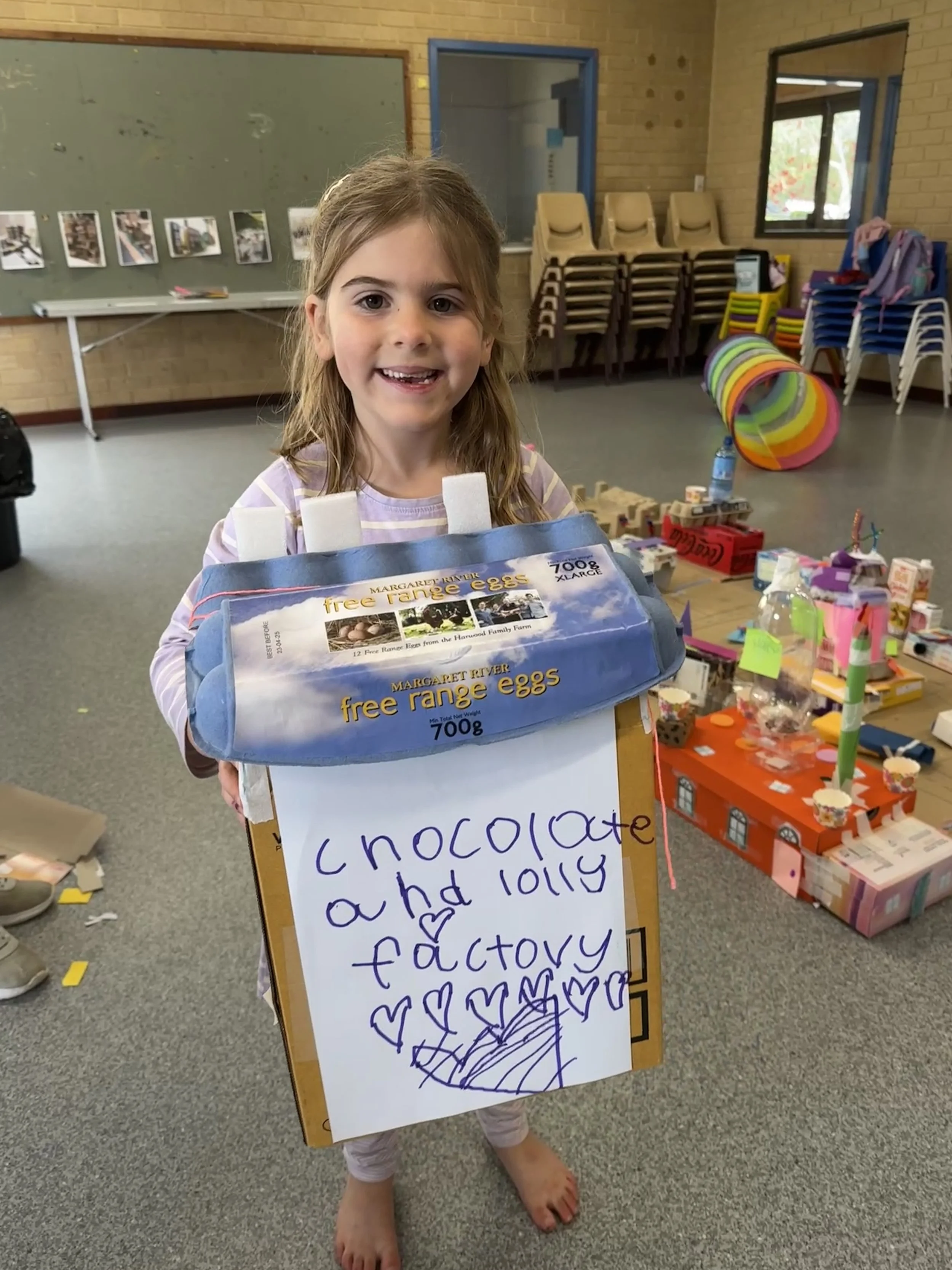 A young girl in pajamas standing indoors in a community center or classroom, smiling and holding a homemade craft project resembling a chocolate factory made from a carton and a food package, with a handwritten sign that says 'Chocolate and lolly fac