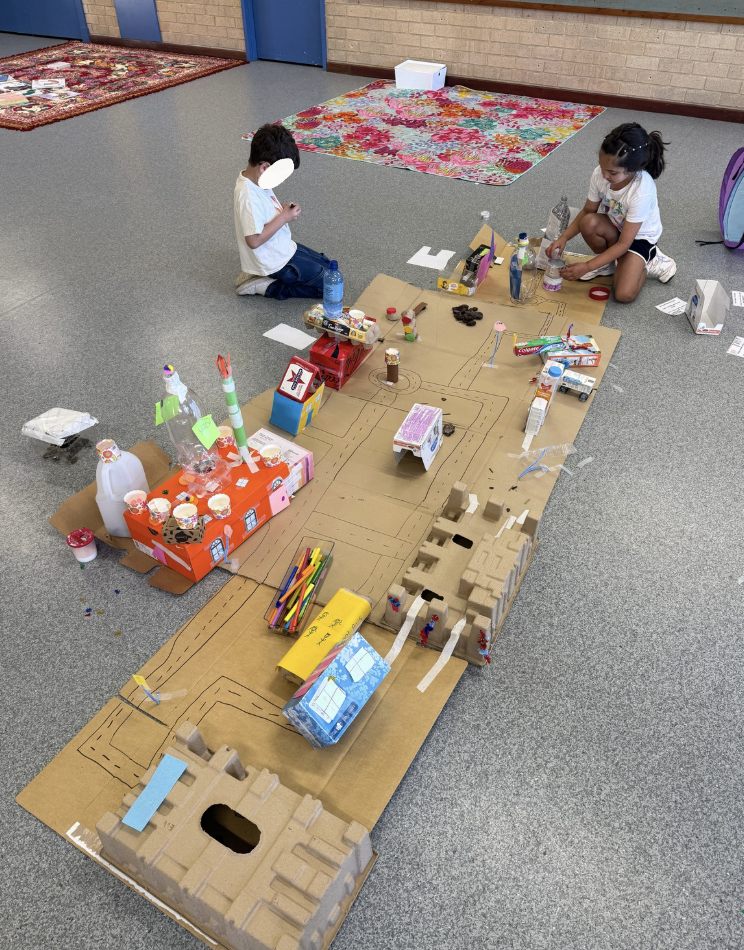 Two children, a boy and a girl, working on a large cardboard city model with various small structures and craft supplies, in a classroom or indoor space.