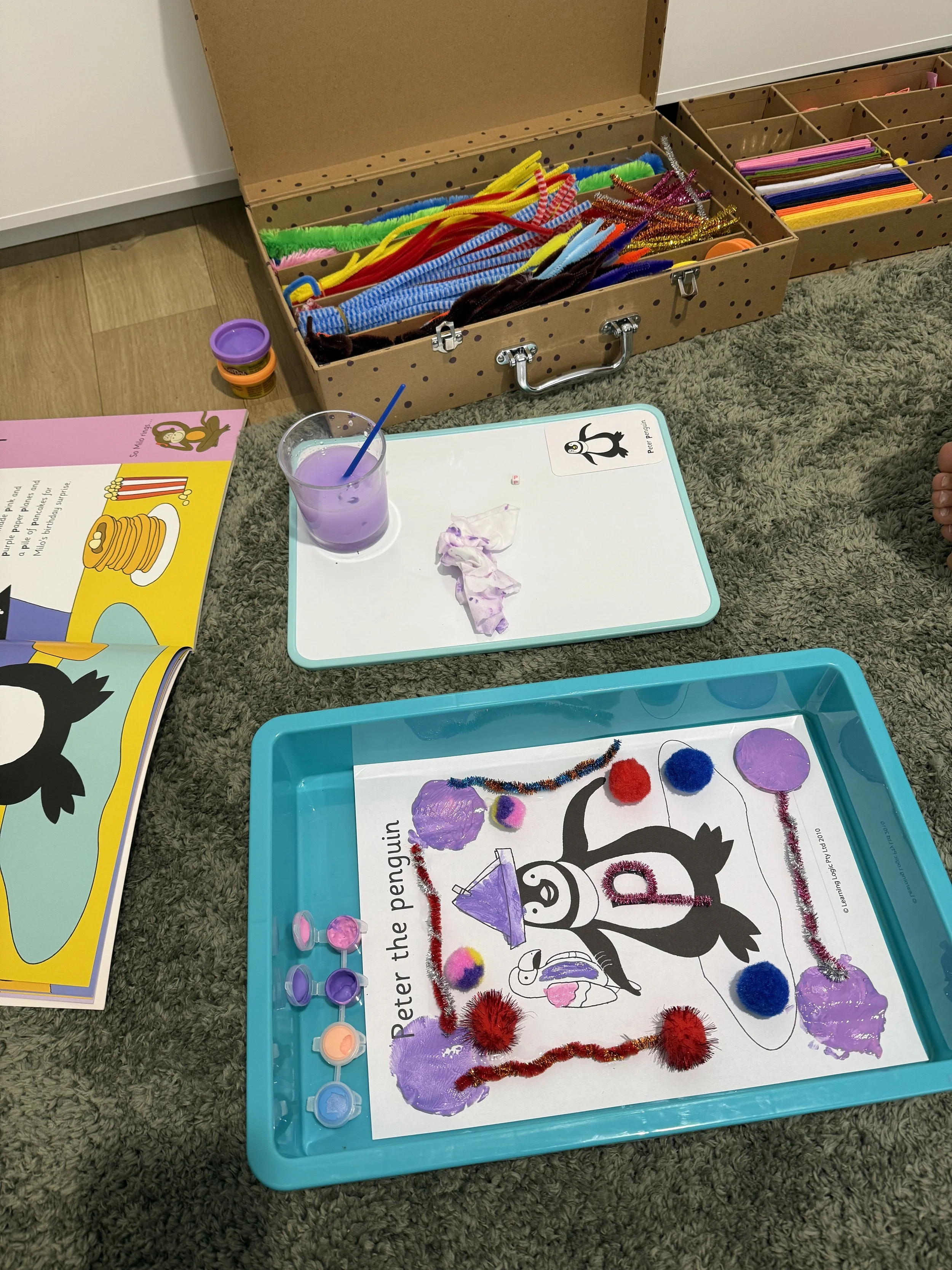A craft activity with a penguin coloring sheet, decorated with purple paint, pom-poms, pipe cleaners, and colored circles on a blue tray. There's a purple drink with a straw, a tissue, a book open to a penguin story, and an organized box of craft supplies in the background.