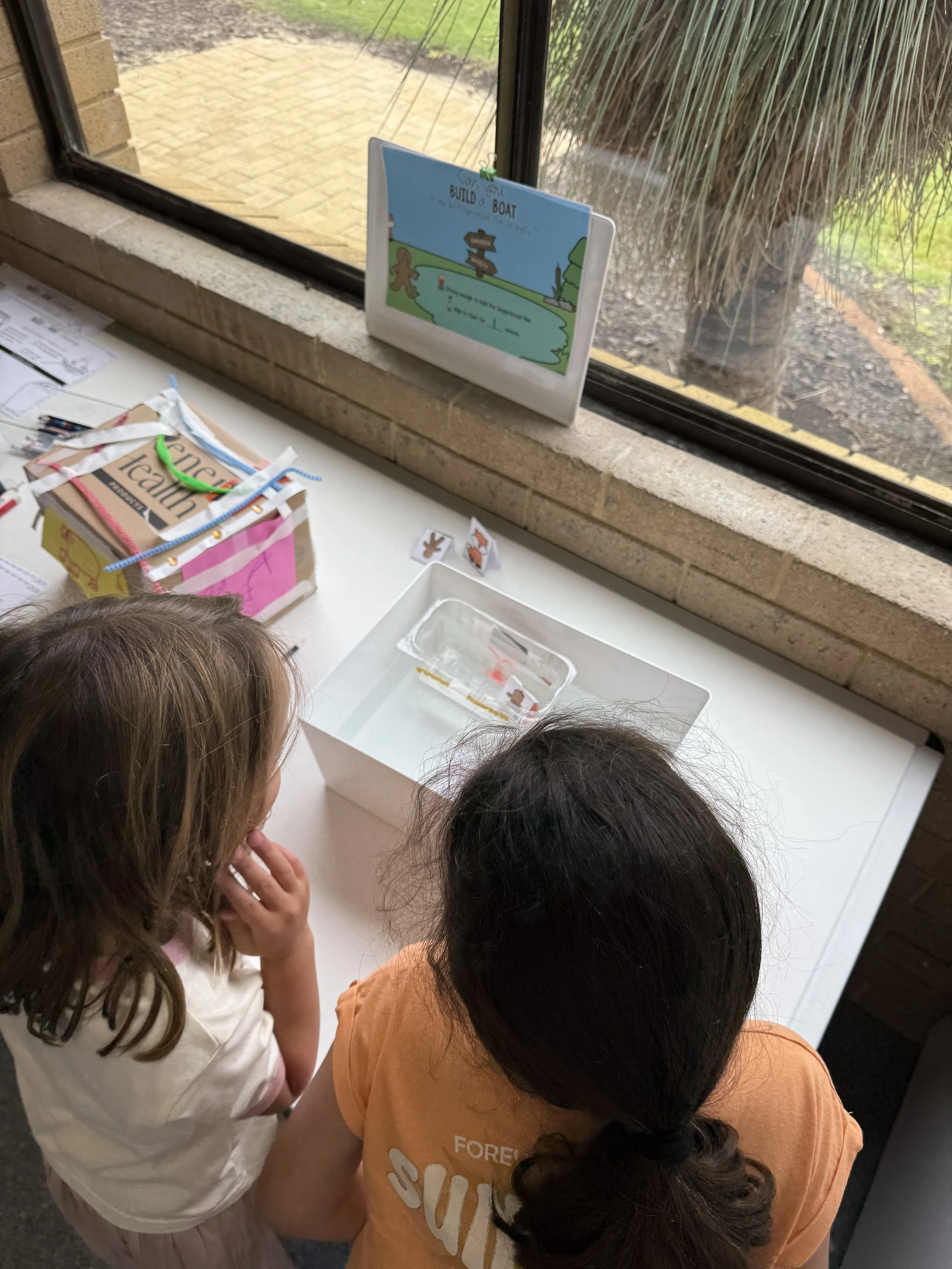 Two children, one with brown hair and one with dark hair, are looking at an activity station on a table near a window, which has a build-a-boat activity and some craft supplies.